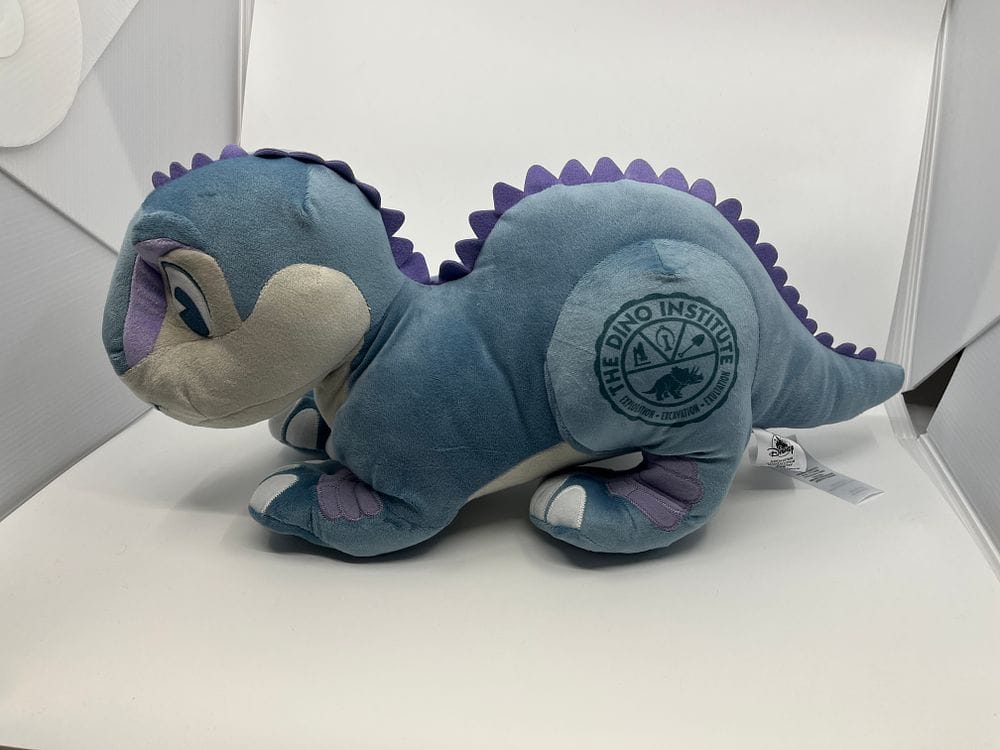 Blue and purple dinosaur plush toy featuring "The Dino Institute" logo on its side, showcased on a white surface. This adorable collectible could make a perfect addition to any collection in the upcoming 2025 WDWNT Spring Cleaning Auction.