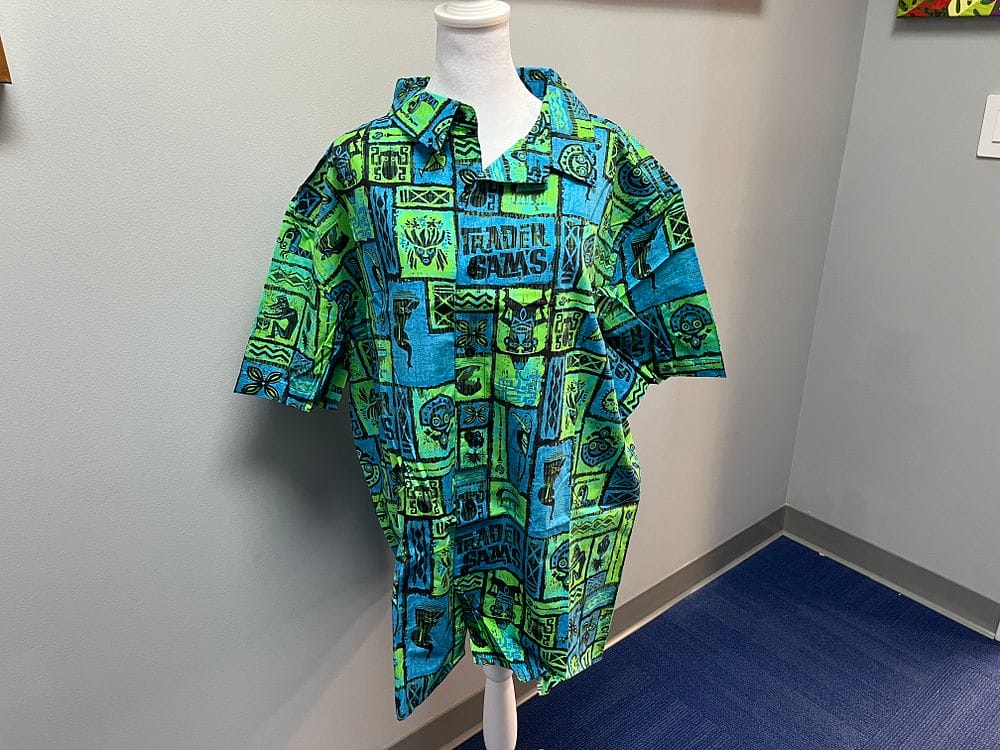 A mannequin dons a vibrant blue and green patterned short-sleeve shirt, showcased in a room with gray walls and a blue carpet, ready to turn heads at the 2025 Spring Cleaning Auction.