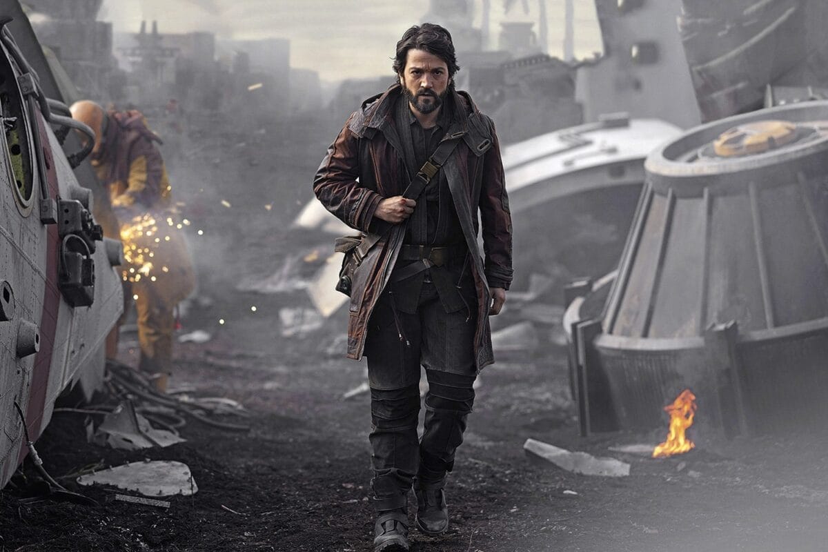 In a dark outfit and brown coat, a man navigates a debris-strewn, smoky landscape. Flames ignite the ground while another individual crafts with sparks in the background—an intense scene perfect for today's Daily Recap.