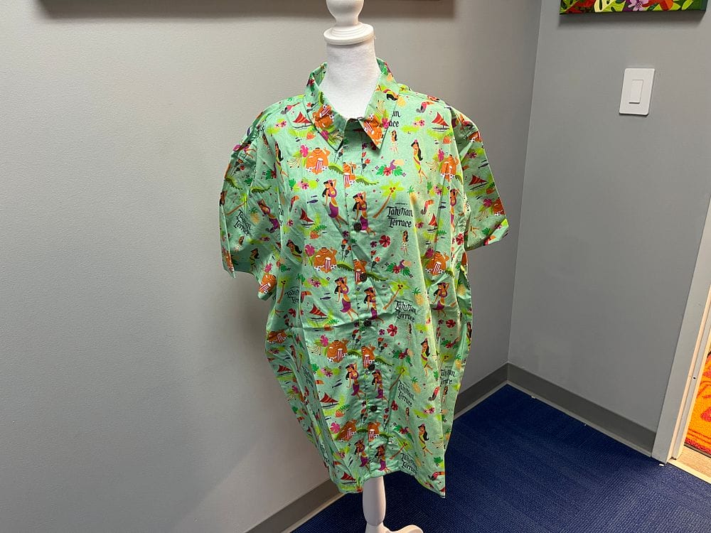 Displayed on a mannequin in a room with gray walls and a blue carpet, this green short-sleeved button-up shirt features colorful cartoon animals. Perfect for those looking to refresh their wardrobes during the 2025 Spring Cleaning Auction.