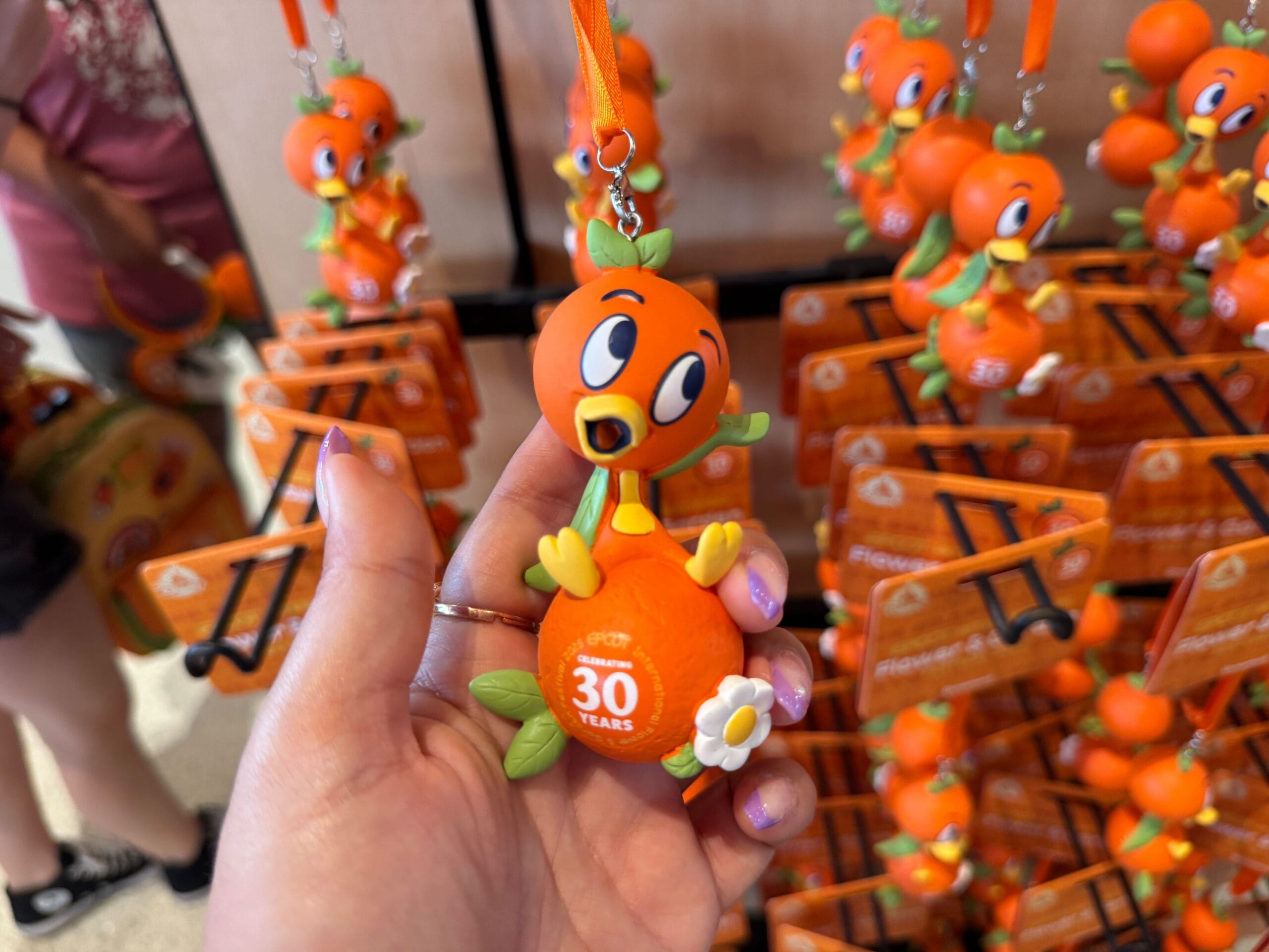 Hand holding a keychain featuring a cartoon orange bird sitting on a 30th-anniversary orange. More keychains in the background.