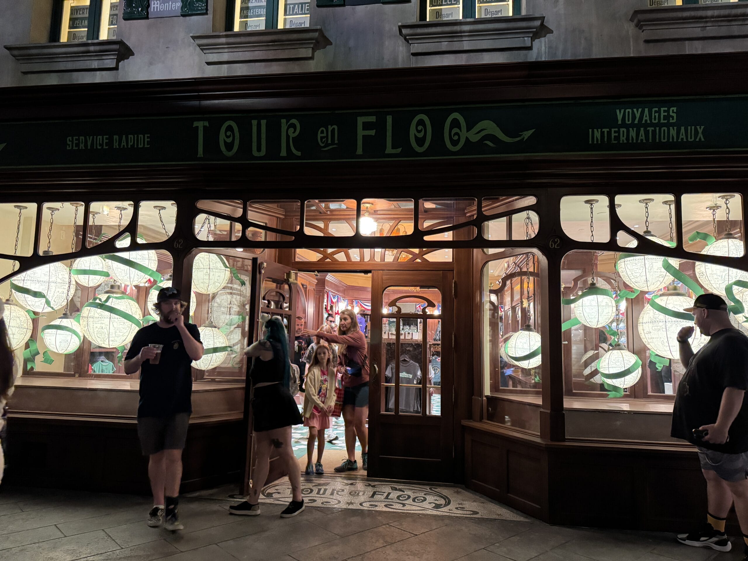 People enter and exit the warmly lit Tour En Floo Shop in the Wizarding World of Harry Potter at Epic Universe at night.