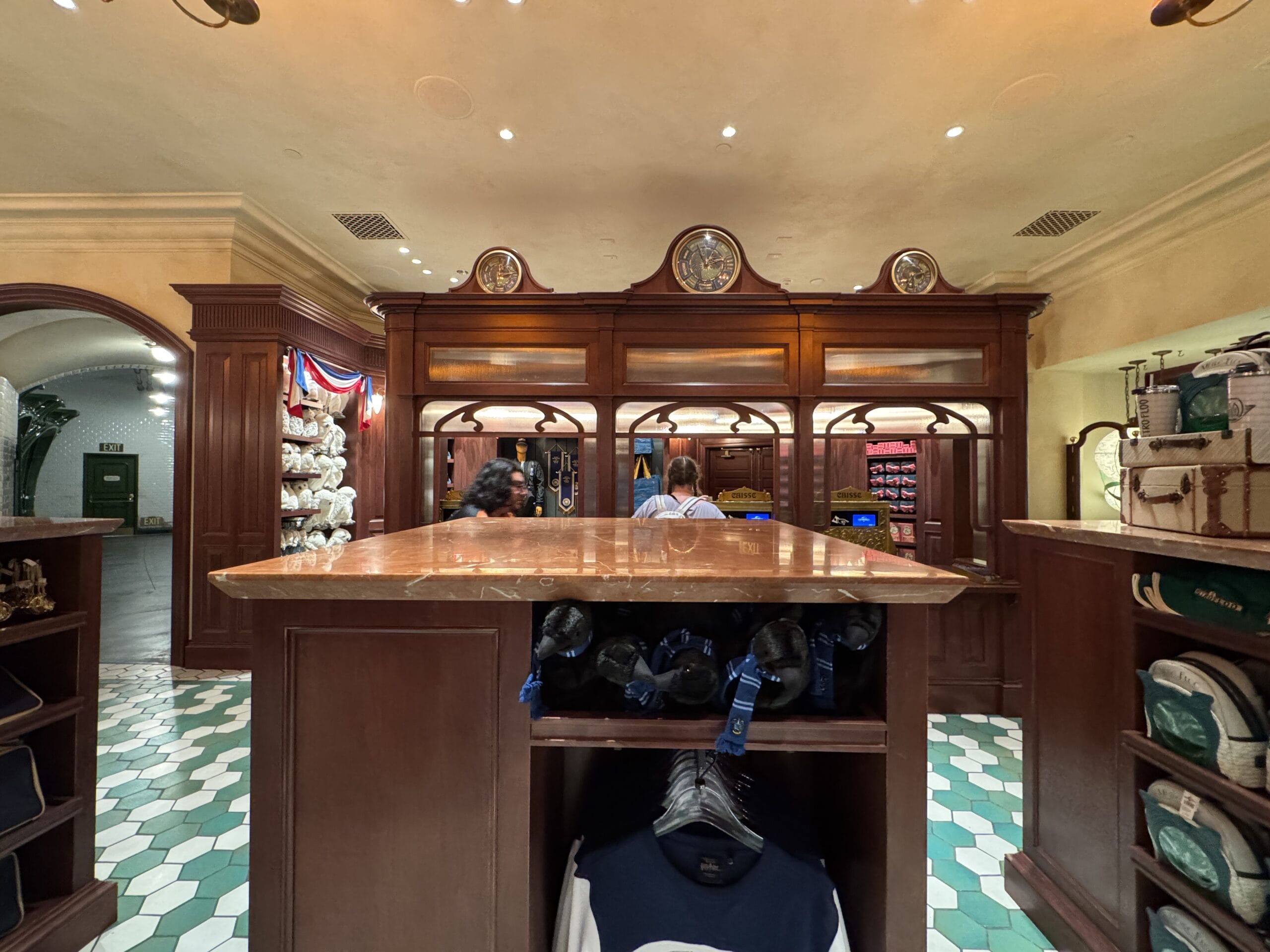 A wooden store counter at the Tour En Floo Shop, with people, shelves of merchandise, and green and white tile floor.