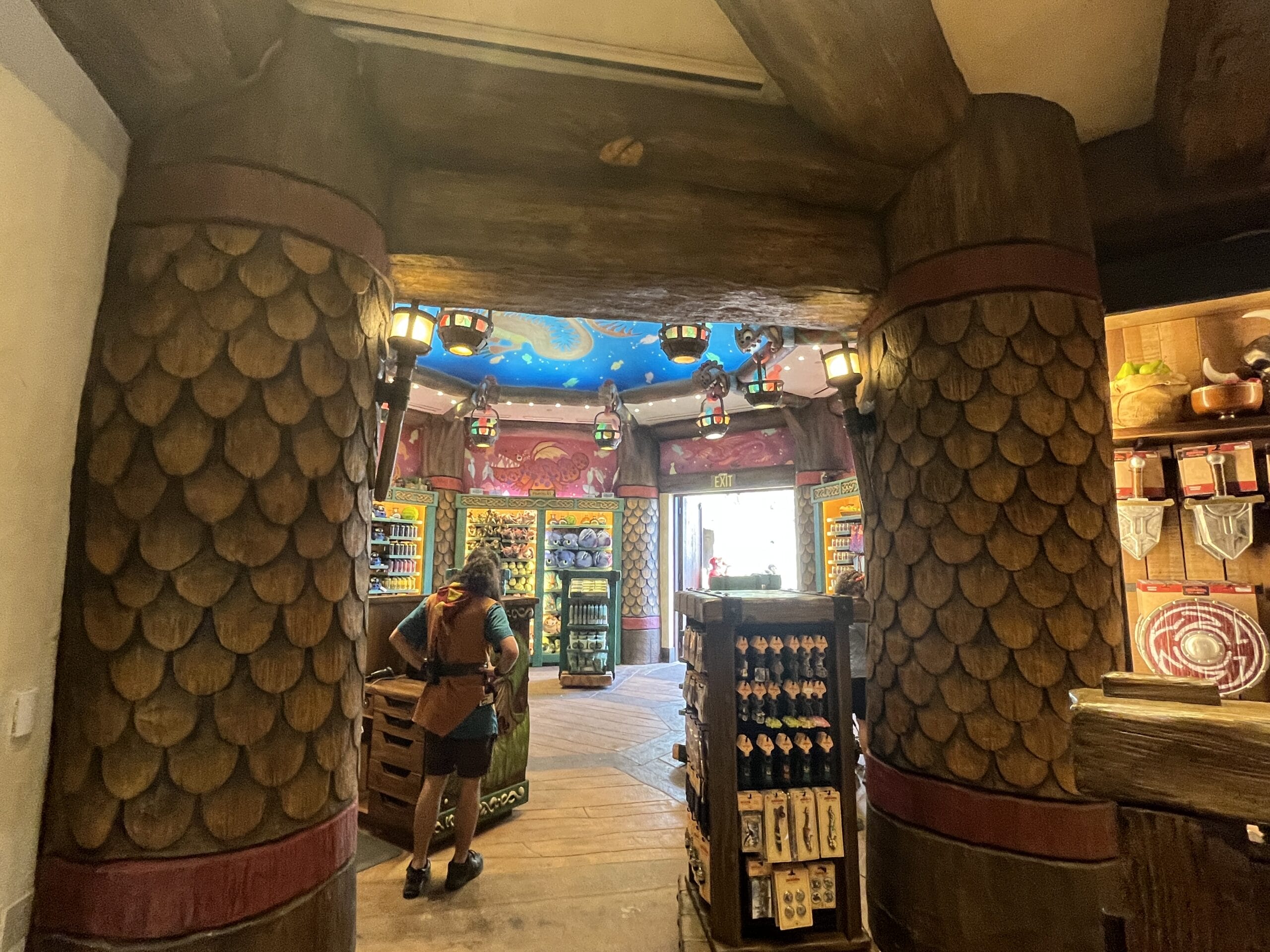 A person stands in Viking Traders, a Berk-themed shop with Nordic wooden decor, lanterns, and vibrant merchandise displays.