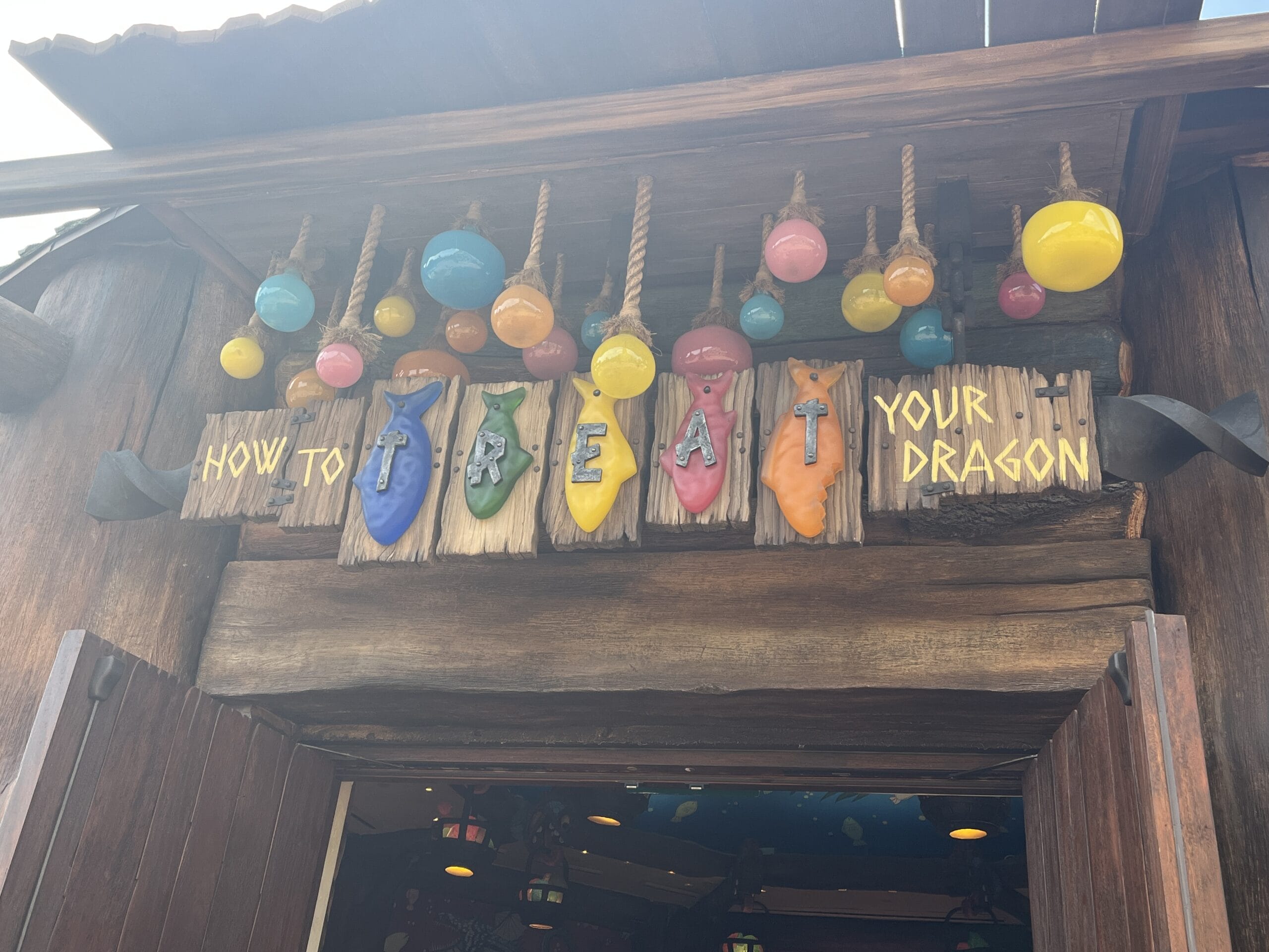 Colorful dragon shapes decorate a wooden sign reading "How to Treat Your Dragon" above a rustic, light-strung doorway.