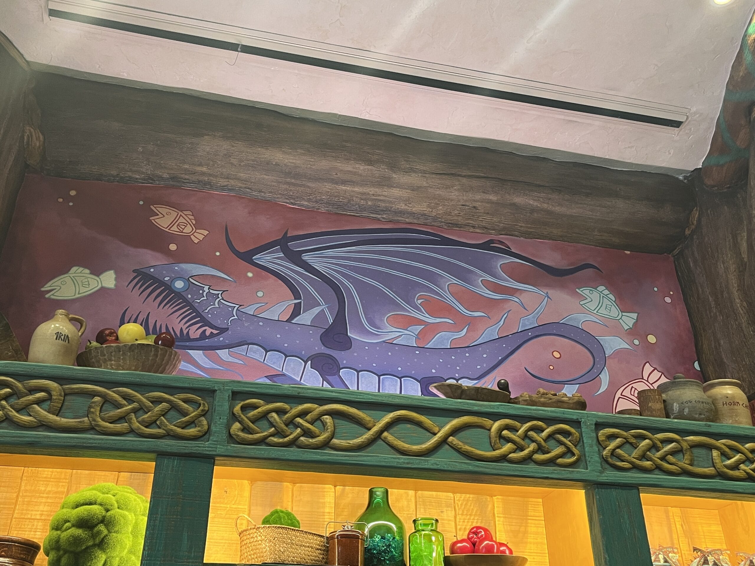 A mural of a dragon with fish, inspired by How to Treat Your Dragon, overlooks jars and food in a Viking-themed room.