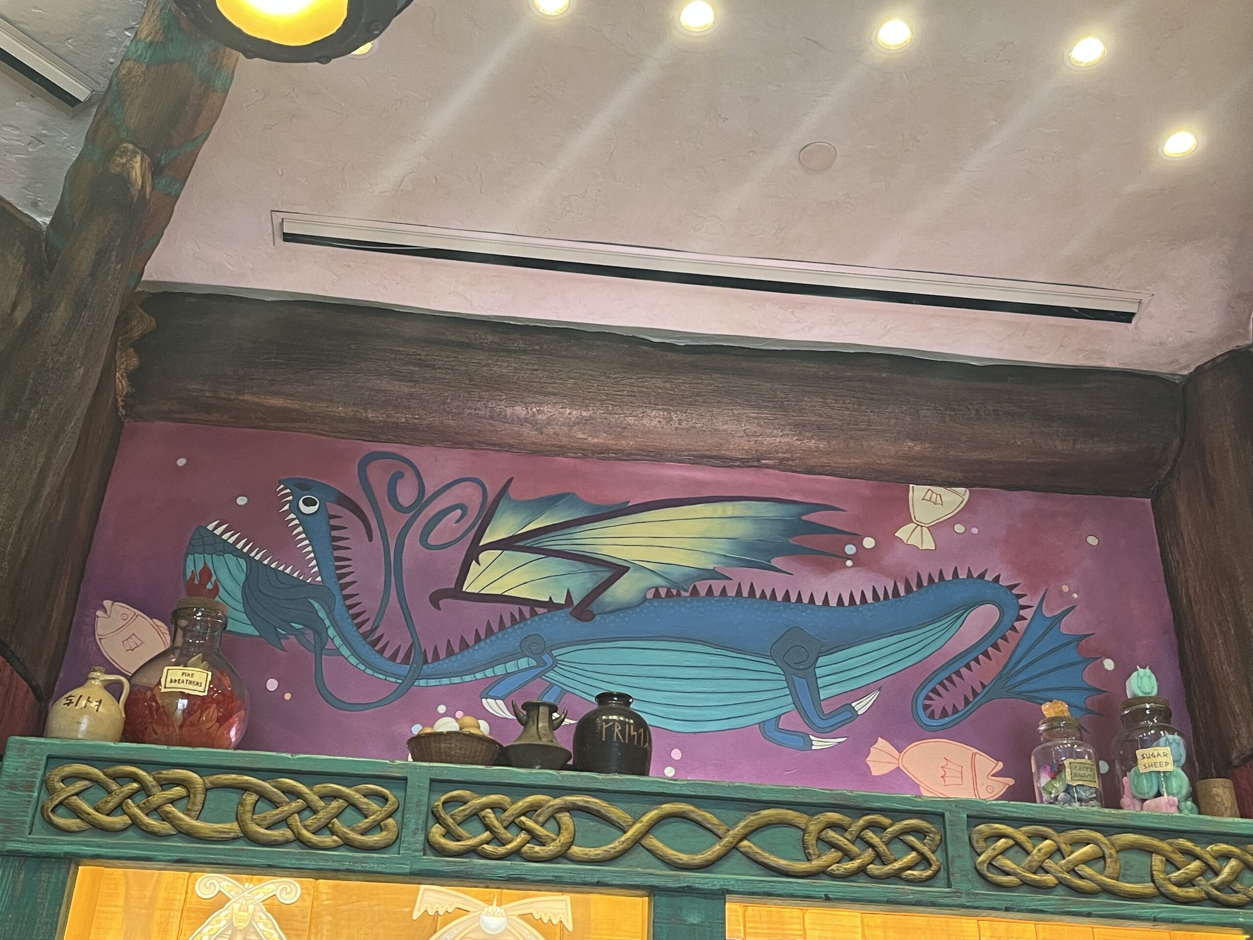 A colorful How to Treat Your Dragon mural above shelves of bottles and jars on a purple wall with Celtic designs.