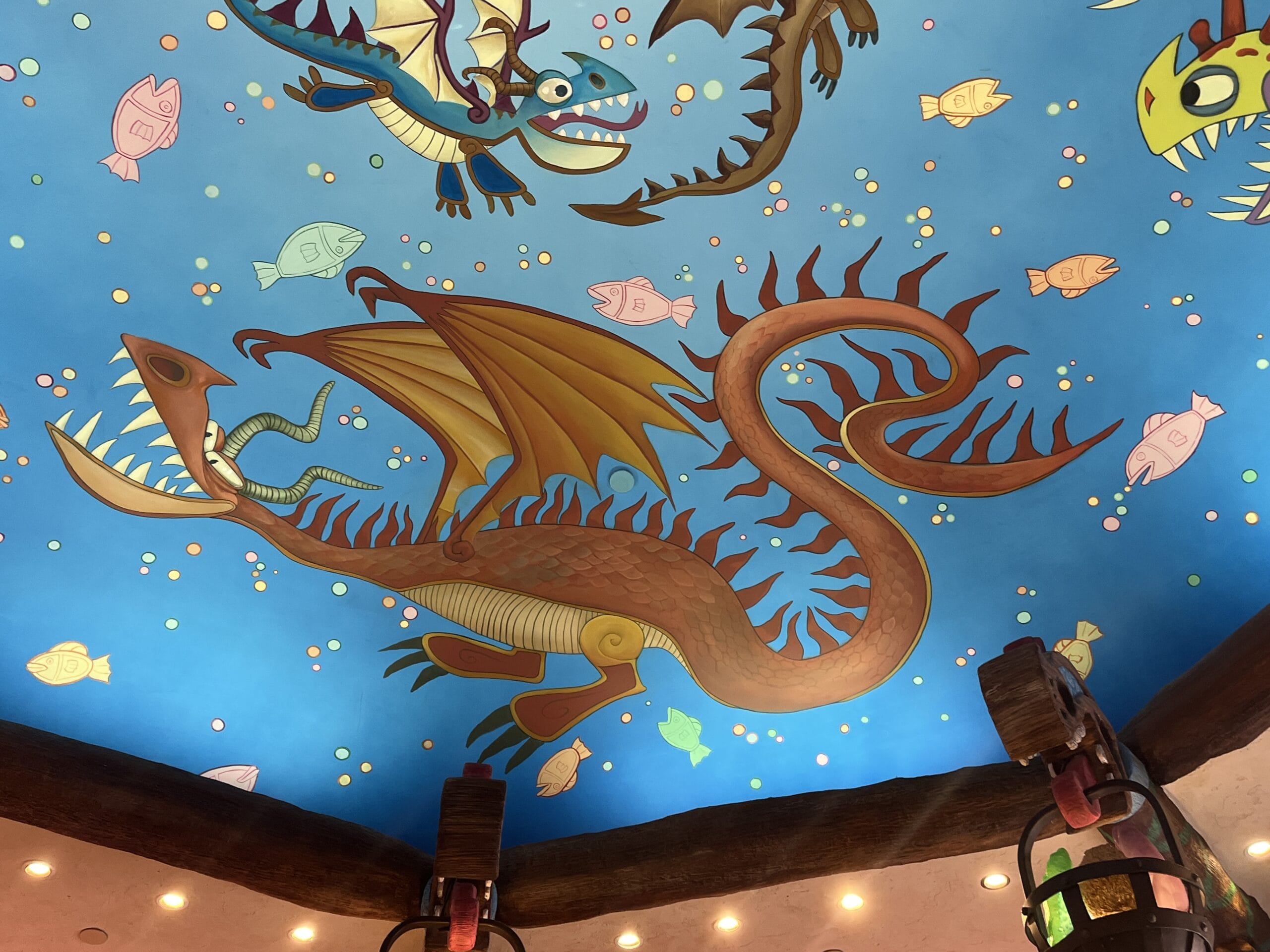 Colorful dragons and fish from How to Treat Your Dragon painted on a blue ceiling, with beams and hanging lanterns below.