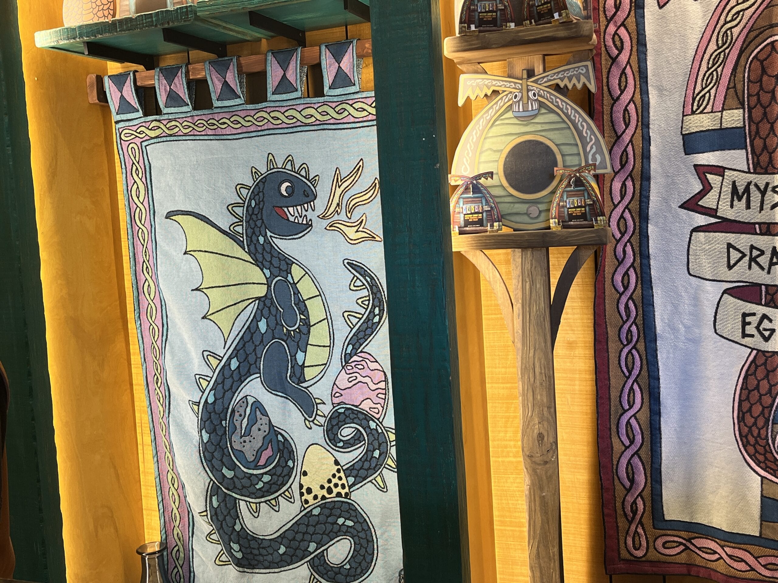Tapestry of a dragon with eggs on a yellow wall, next to Viking mugs—perfect for How to Treat Your Dragon fans.