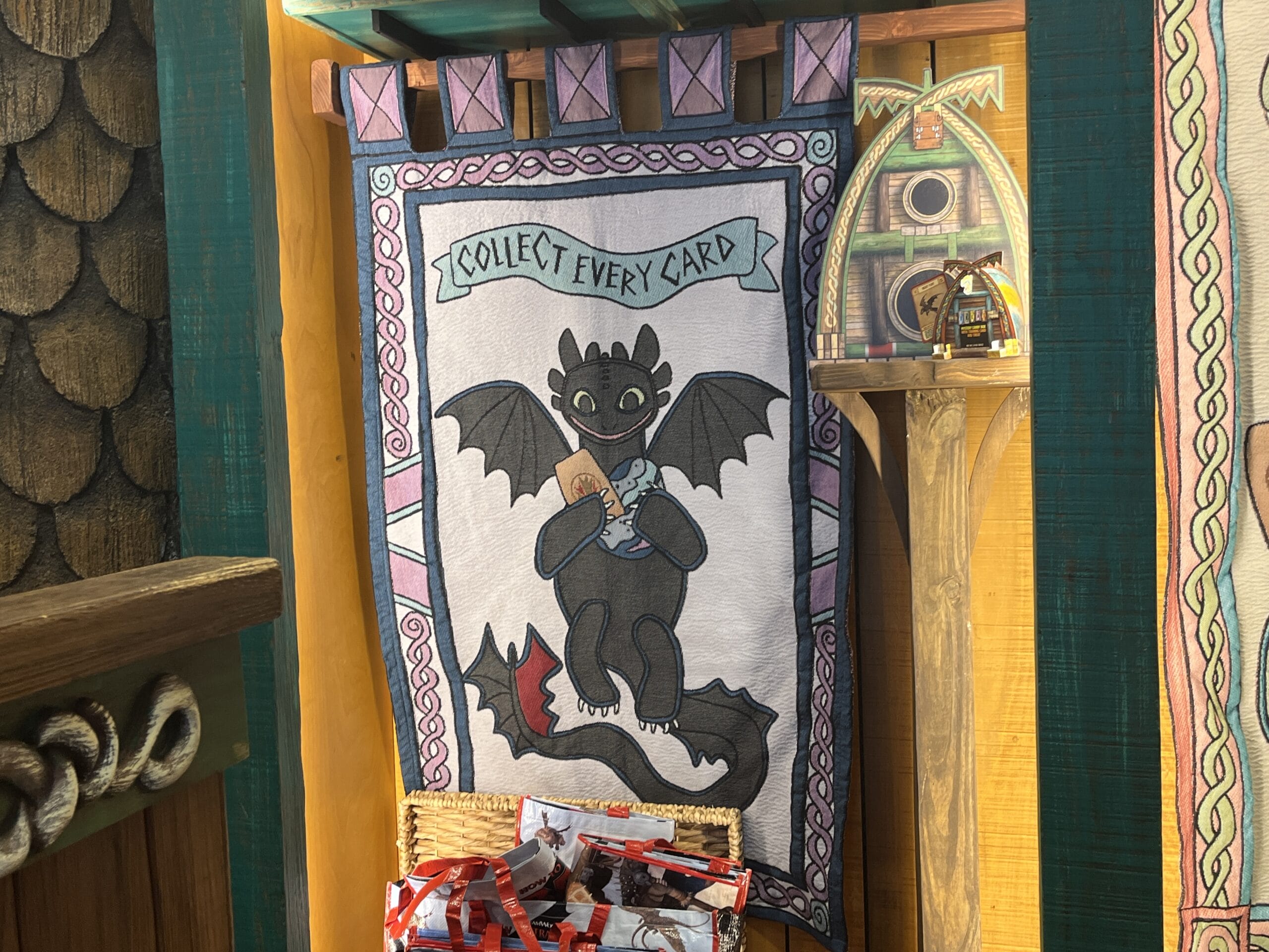 A cartoon dragon from How to Treat Your Dragon holds a card on a tapestry that says "Collect Every Card.
