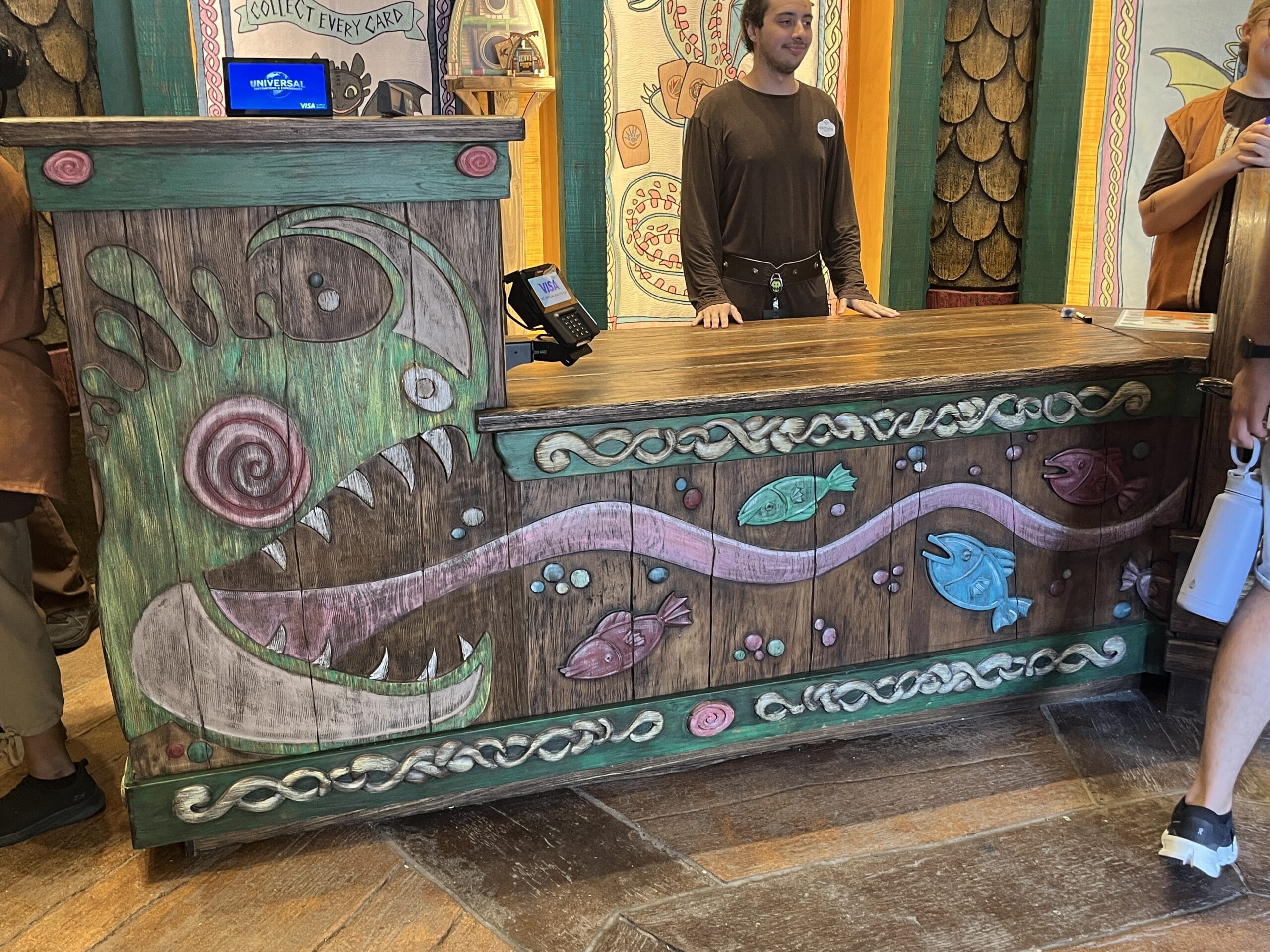A wooden counter with colorful fish and How to Treat Your Dragon sea creature art, with people standing nearby.