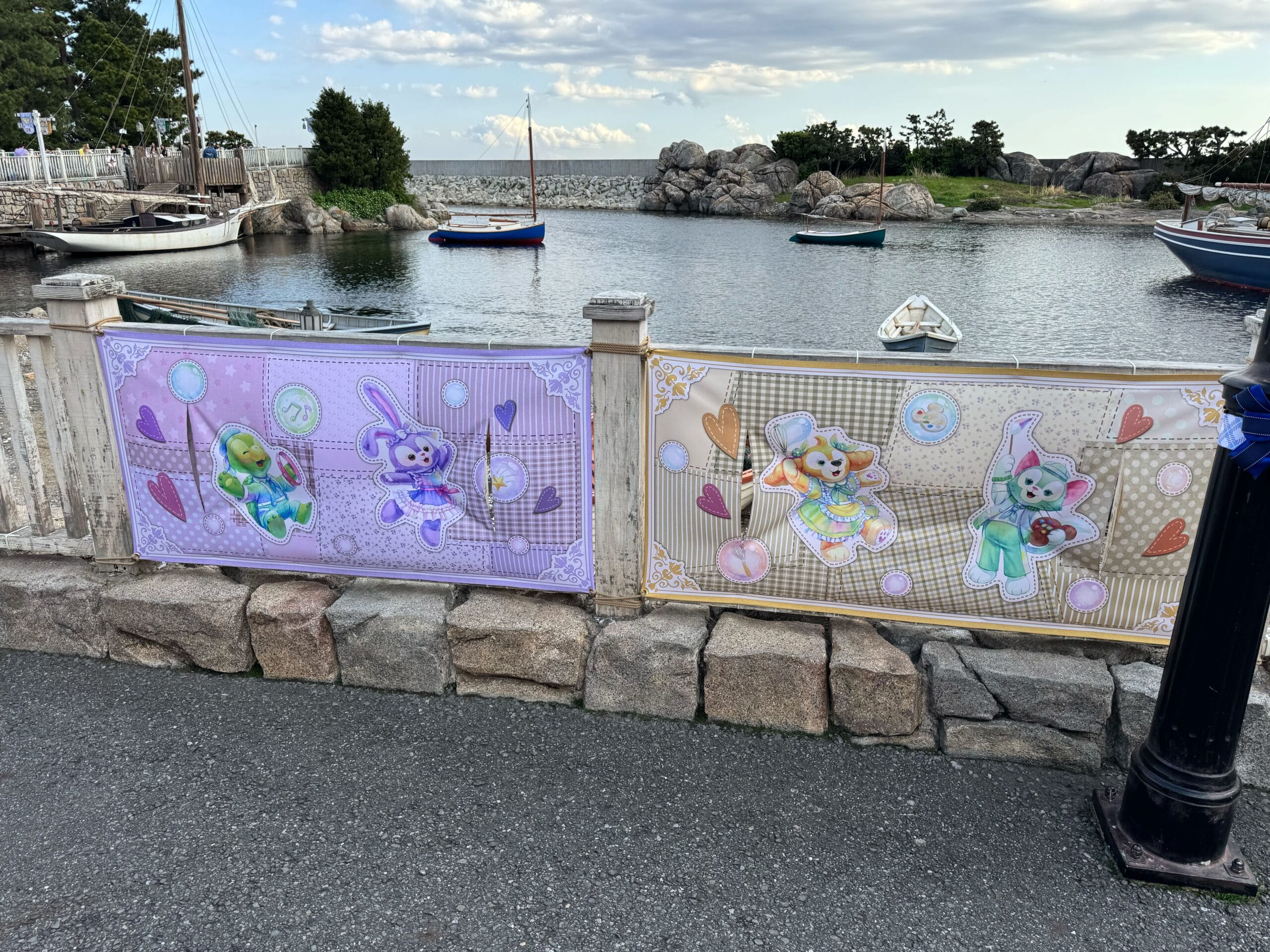 Two illustrated banners featuring Duffy and Friends' pastel-colored bears and rabbits are attached to a white fence by the waterfront at Tokyo DisneySea, with boats and rocks in the background.