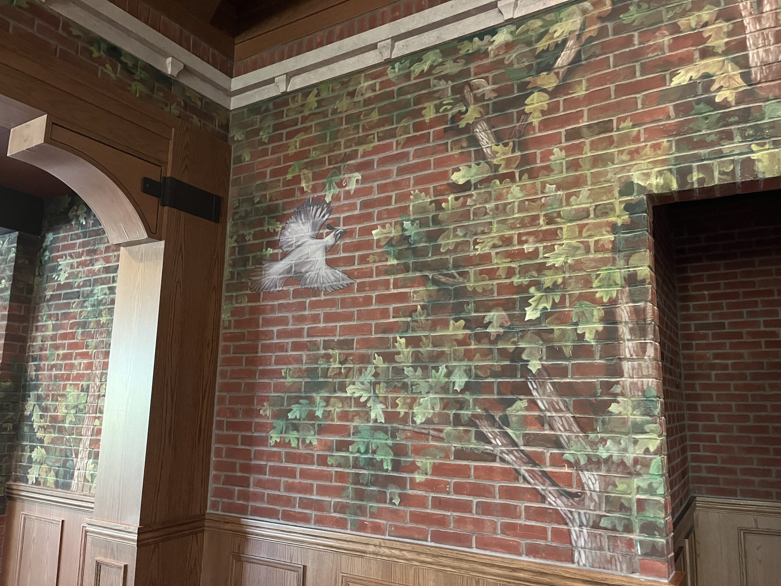 A mural of a bird and green leaves on a red brick wall at The Oak & Star Tavern, above wood paneling, reminiscent of Disney park artistry.
