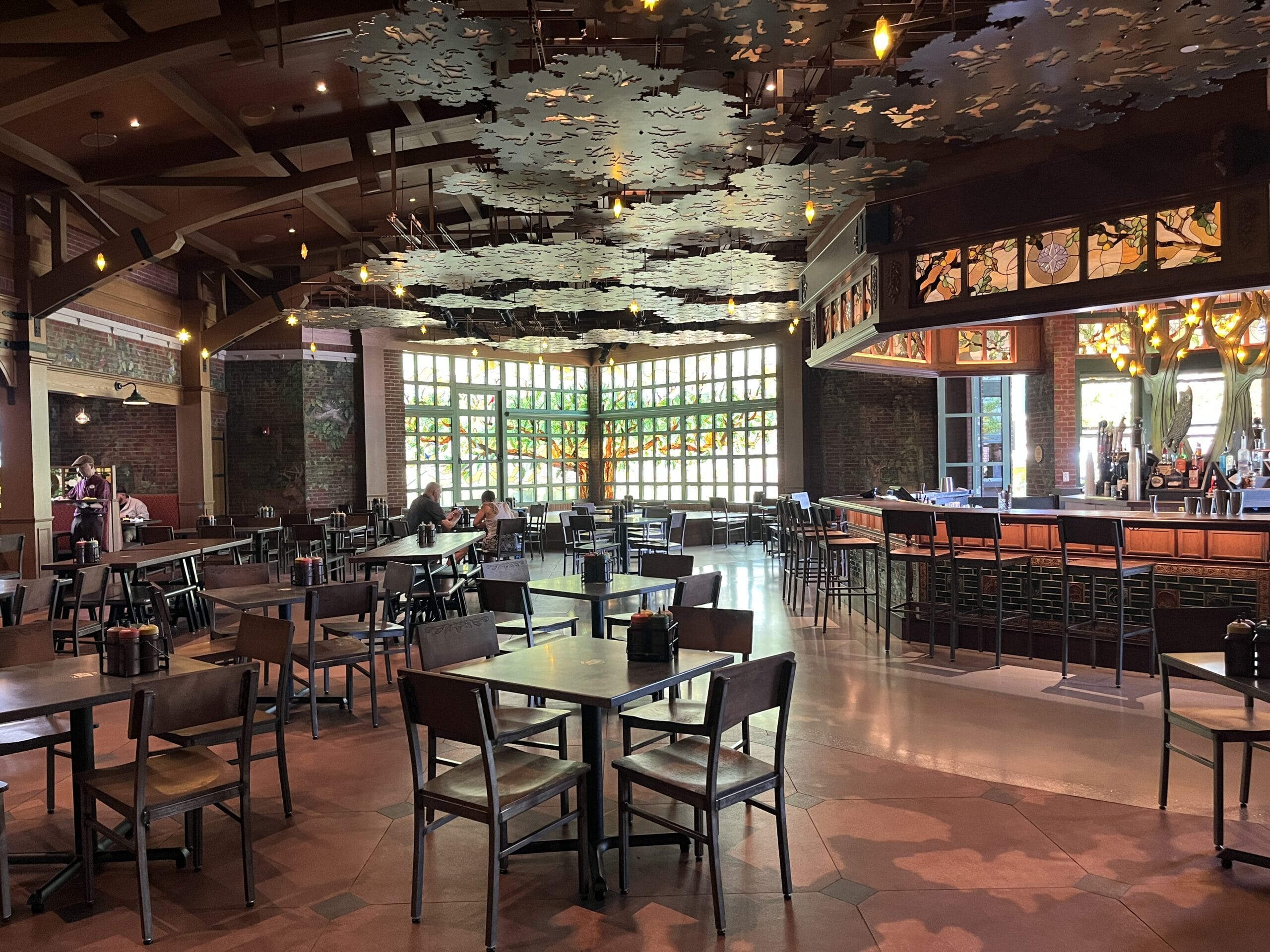 The Oak & Star Tavern, a roomy Disney park restaurant with wooden tables, large windows, unique ceiling art, and a bar on the right.