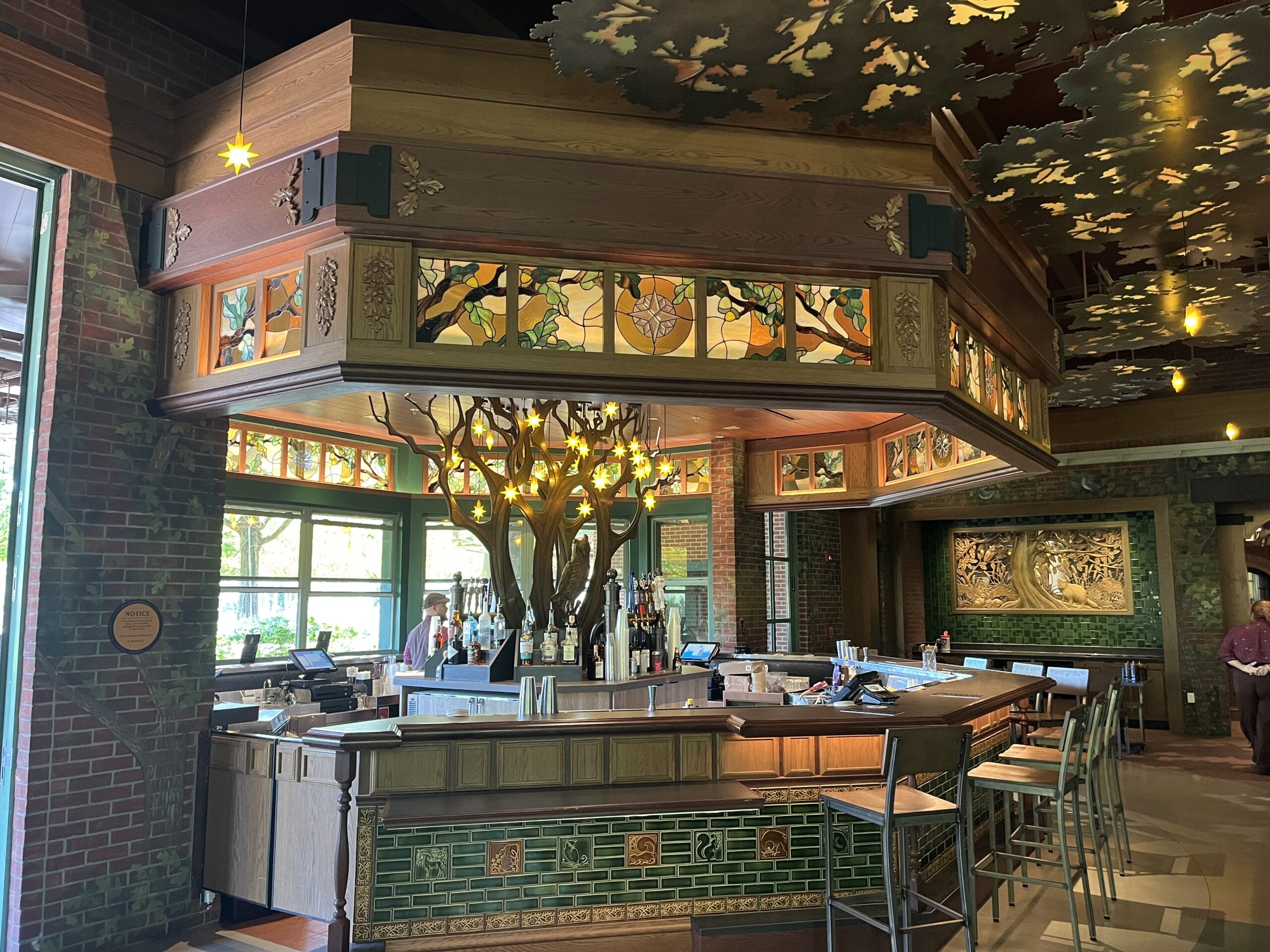 The Oak & Star Tavern, in a cozy Disney park style, displays stained glass, green tiles, and a whimsical tree-like light fixture.