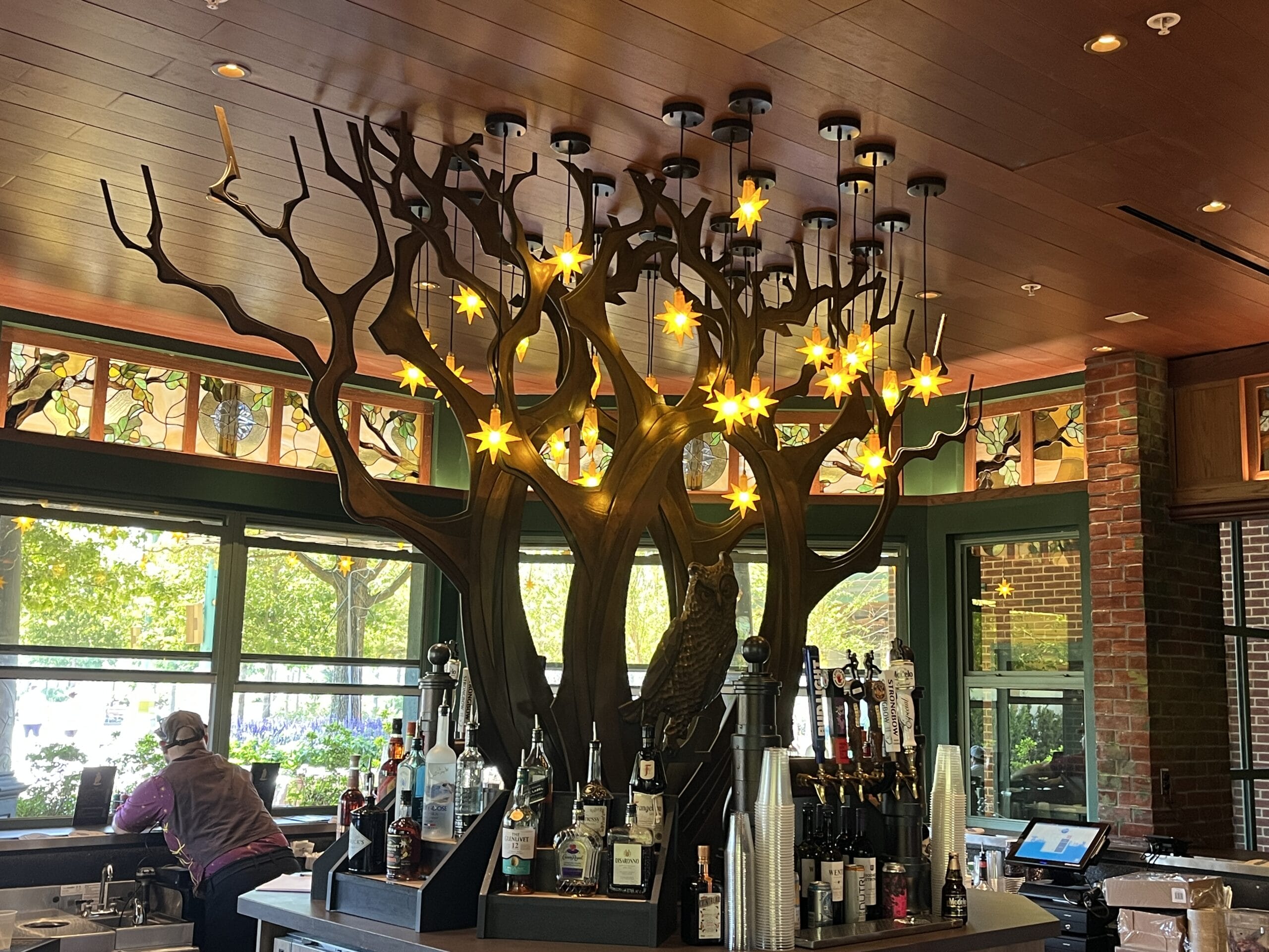 A guest stands at The Oak & Star Tavern’s bar, beneath whimsical tree-shaped lights and star lamps, greenery visible outside—evoking the immersive theming often found at Walt Disney Parks lounges.
