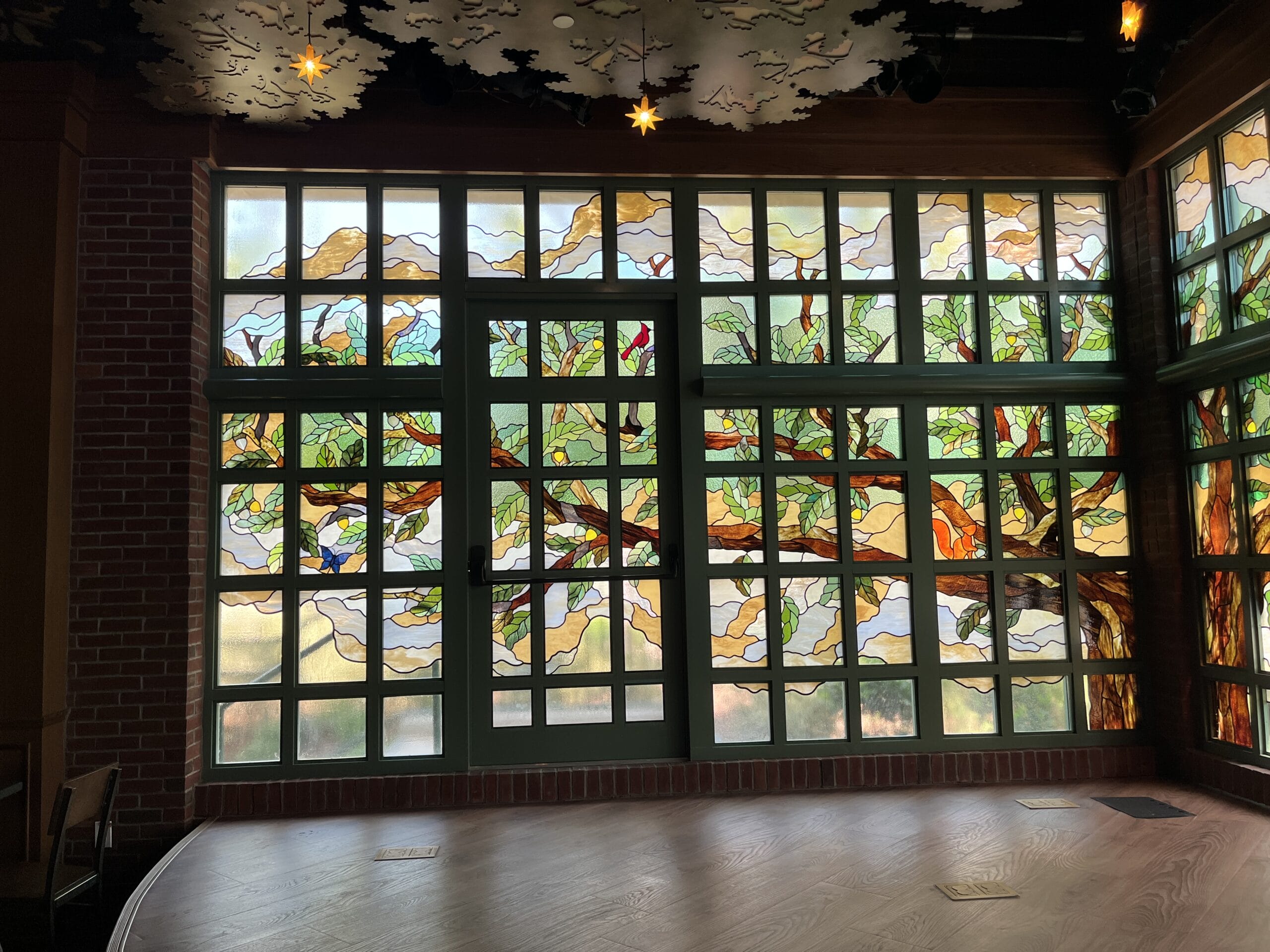 Stained glass windows with a vibrant tree, leaves, and birds illuminate The Oak & Star Tavern’s stage like Disney park magic.