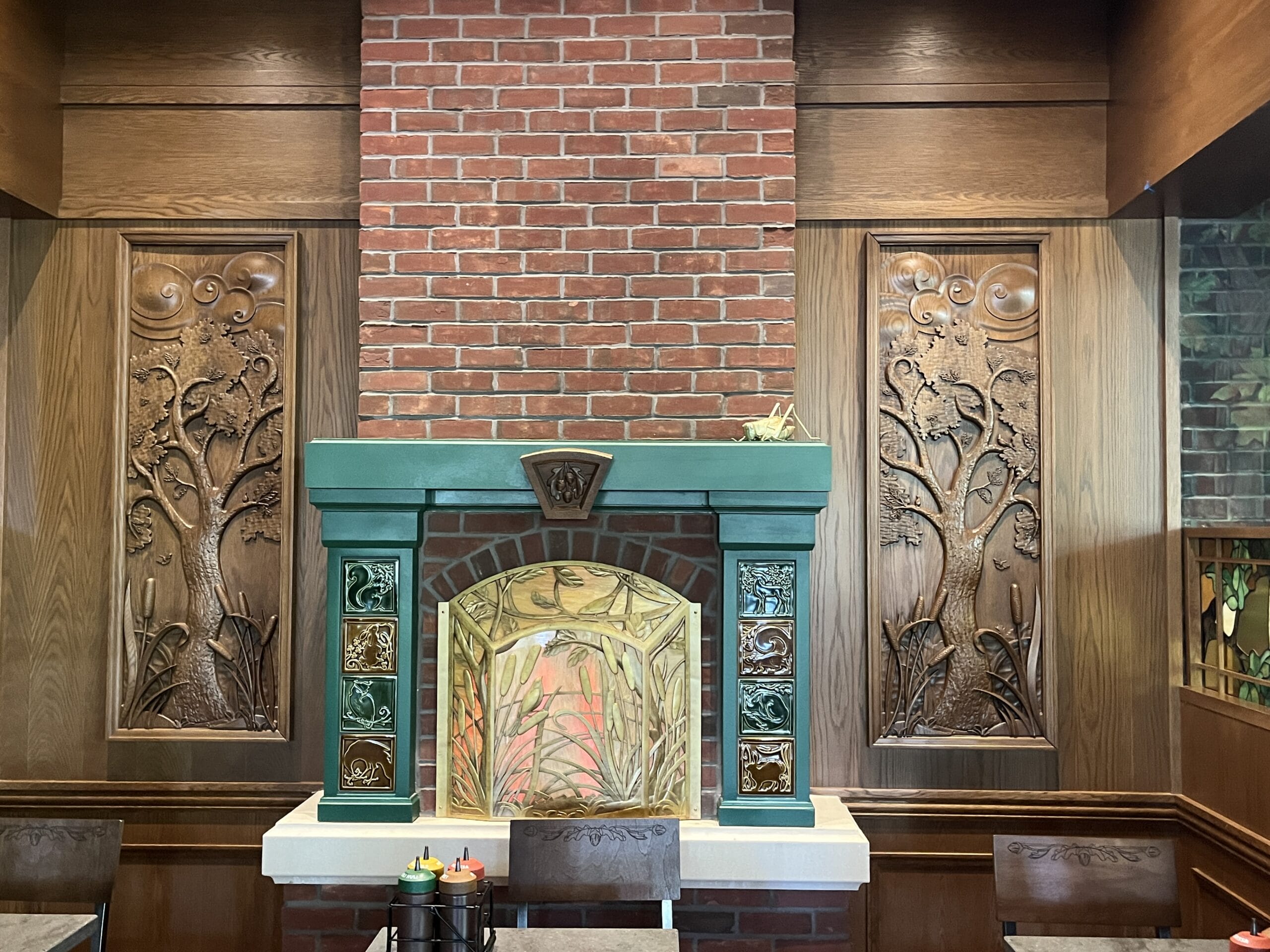 Brick fireplace with green and gold tiles at The Oak & Star Tavern, framed by wood panels carved with tree motifs, like Disney decor.