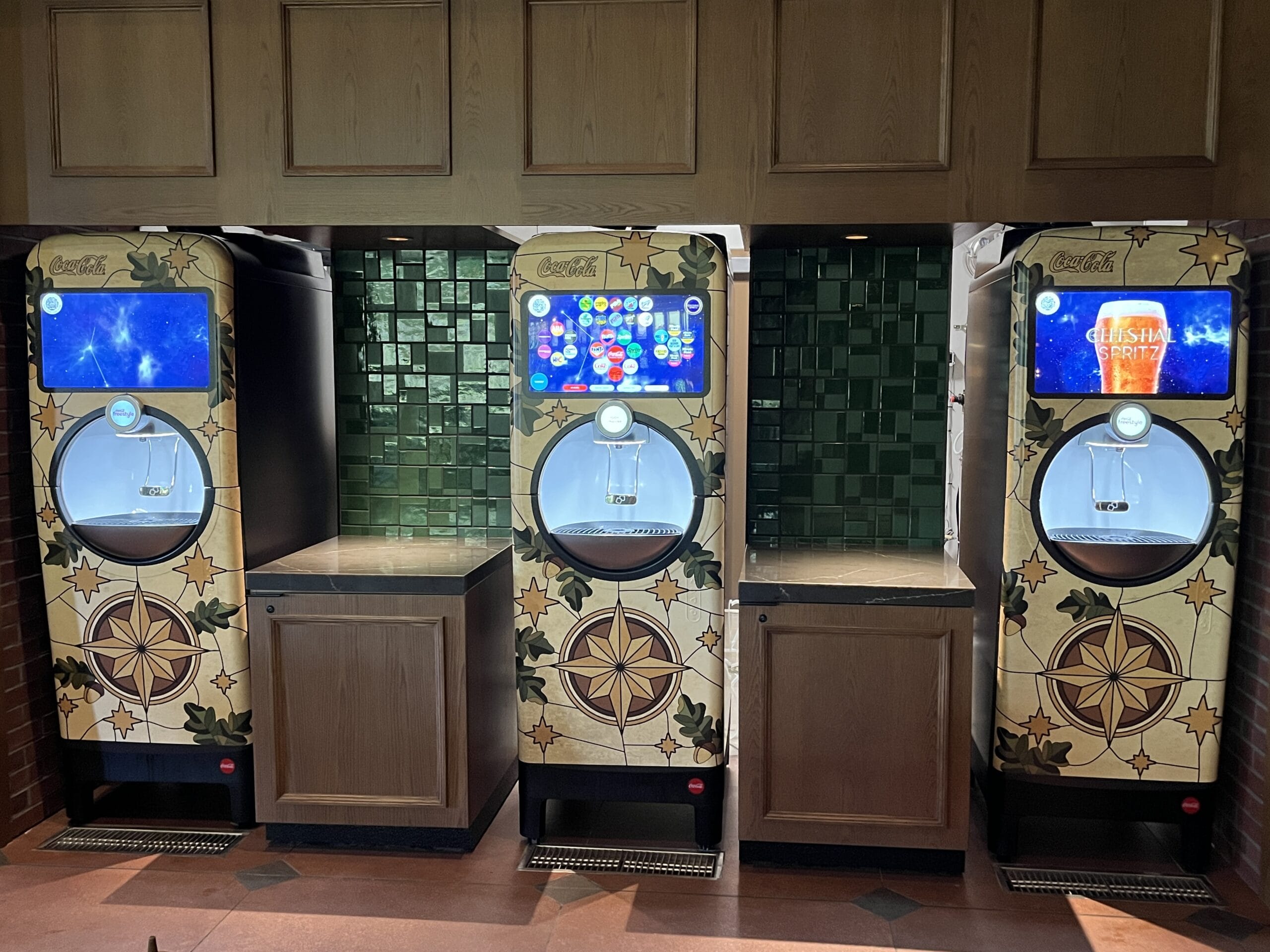 Three Coca-Cola freestyle machines at The Oak & Star Tavern, decorative panels and green tiles evoke whimsical Disney park theming.