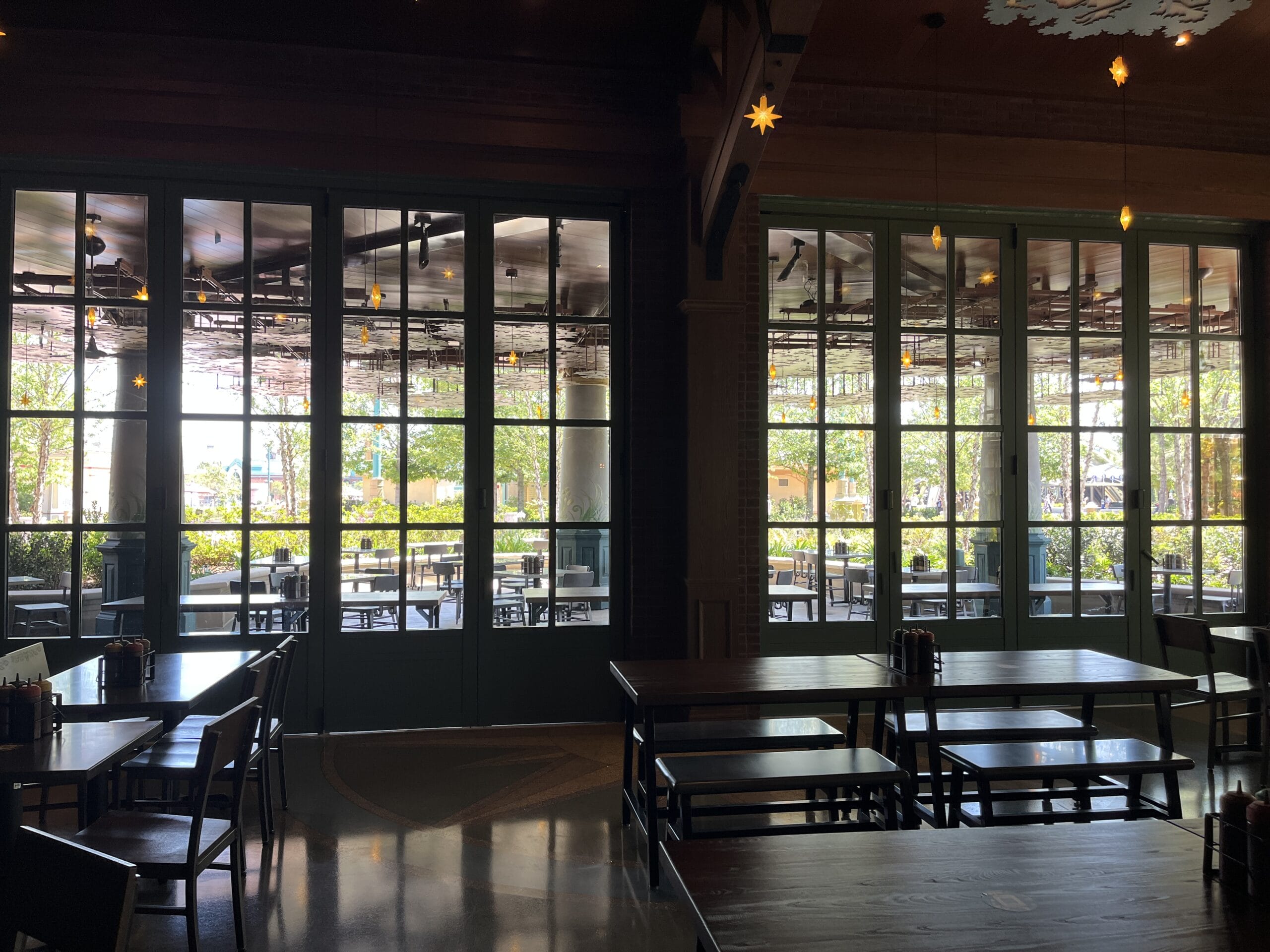 Cozy, dim interior of The Oak & Star Tavern; empty tables overlook sunny patio and lush greenery, reminiscent of Epcot lounges.