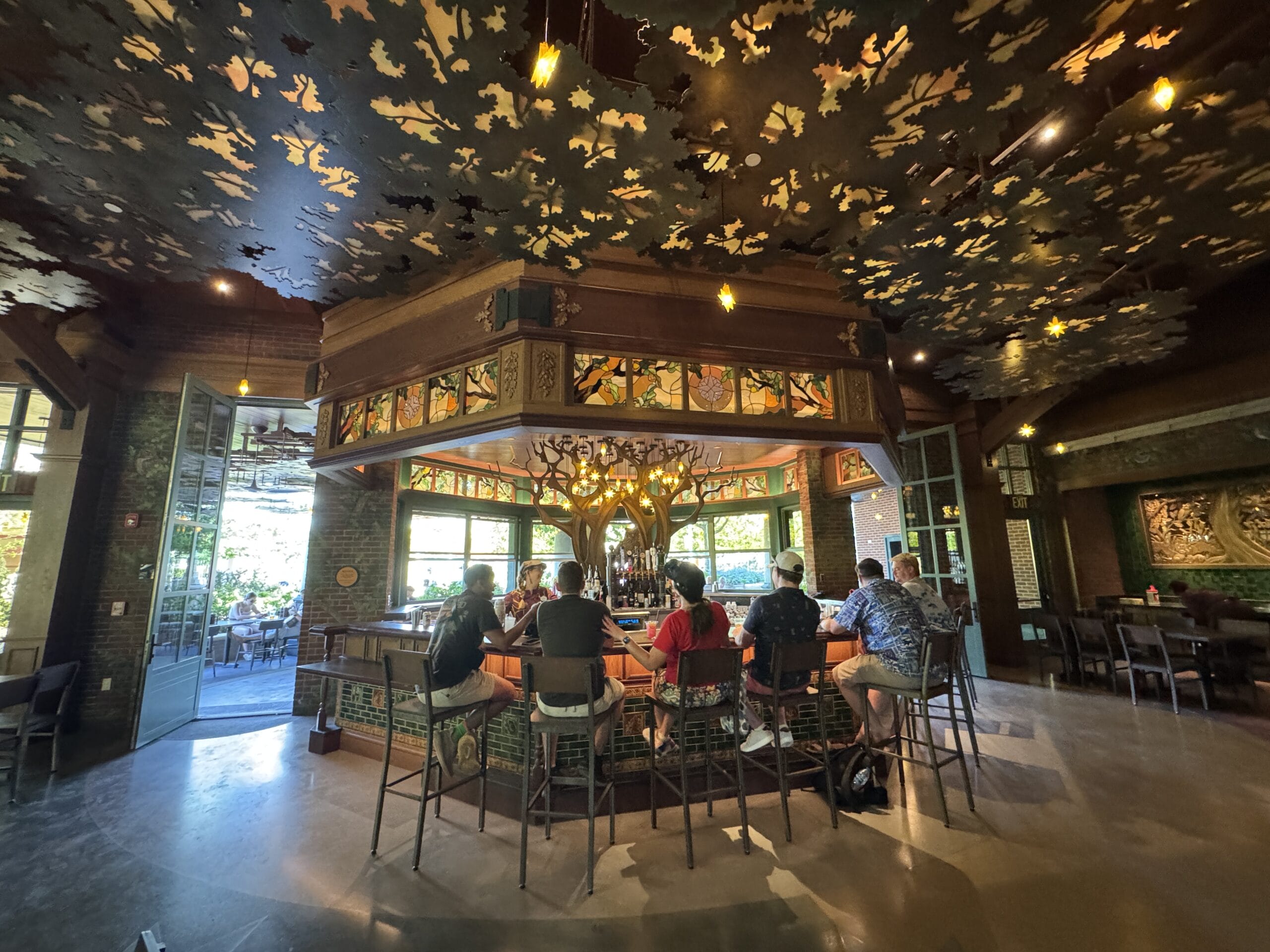 Guests gather at a cozy bar in The Oak & Star Tavern, its stained glass and ornate ceiling echoing classic Disney park ambiance.