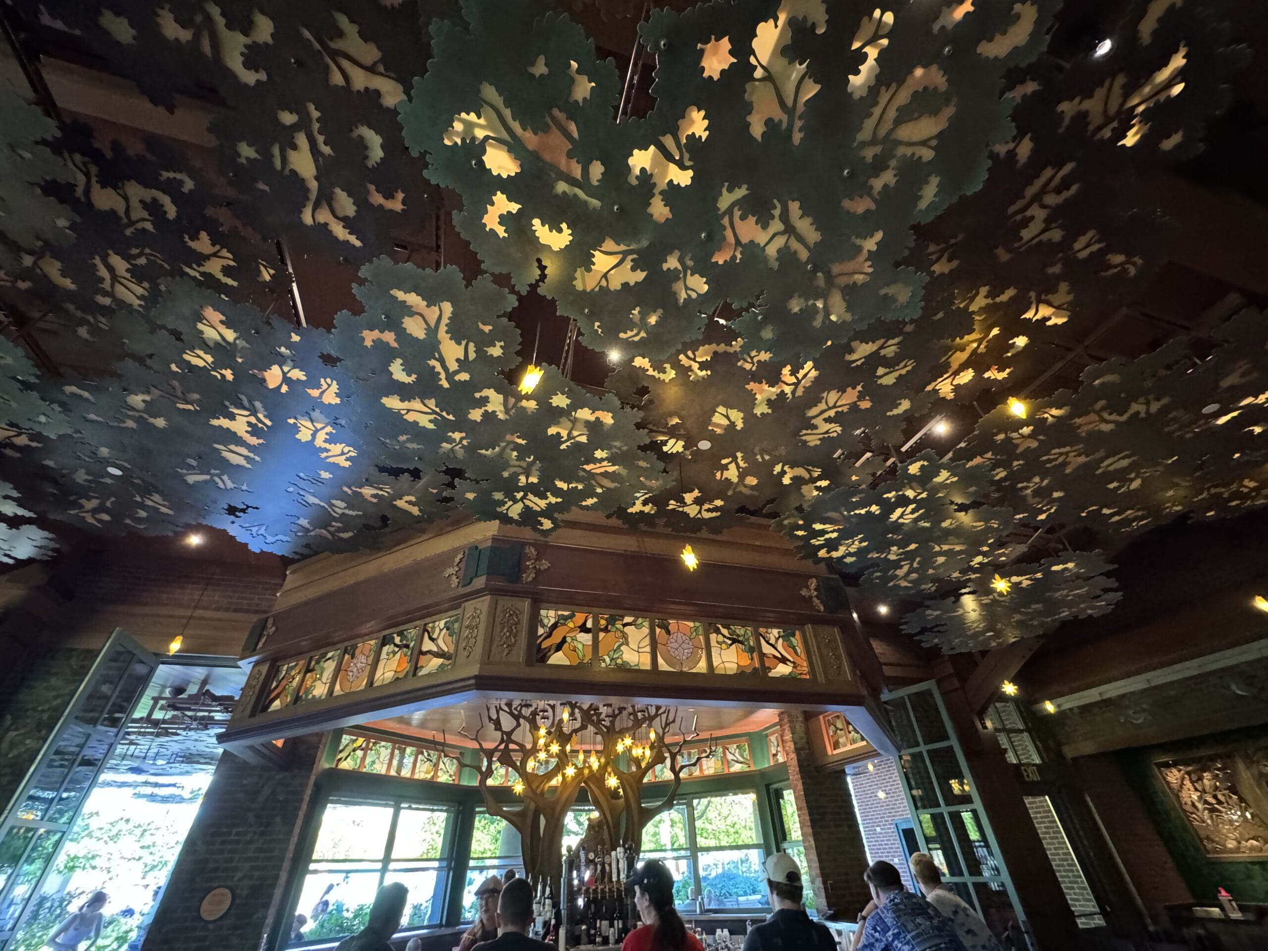 Inside The Oak & Star Tavern, guests dine beneath a leafy, enchanted ceiling. Large windows offer views, much like Disney park eateries.