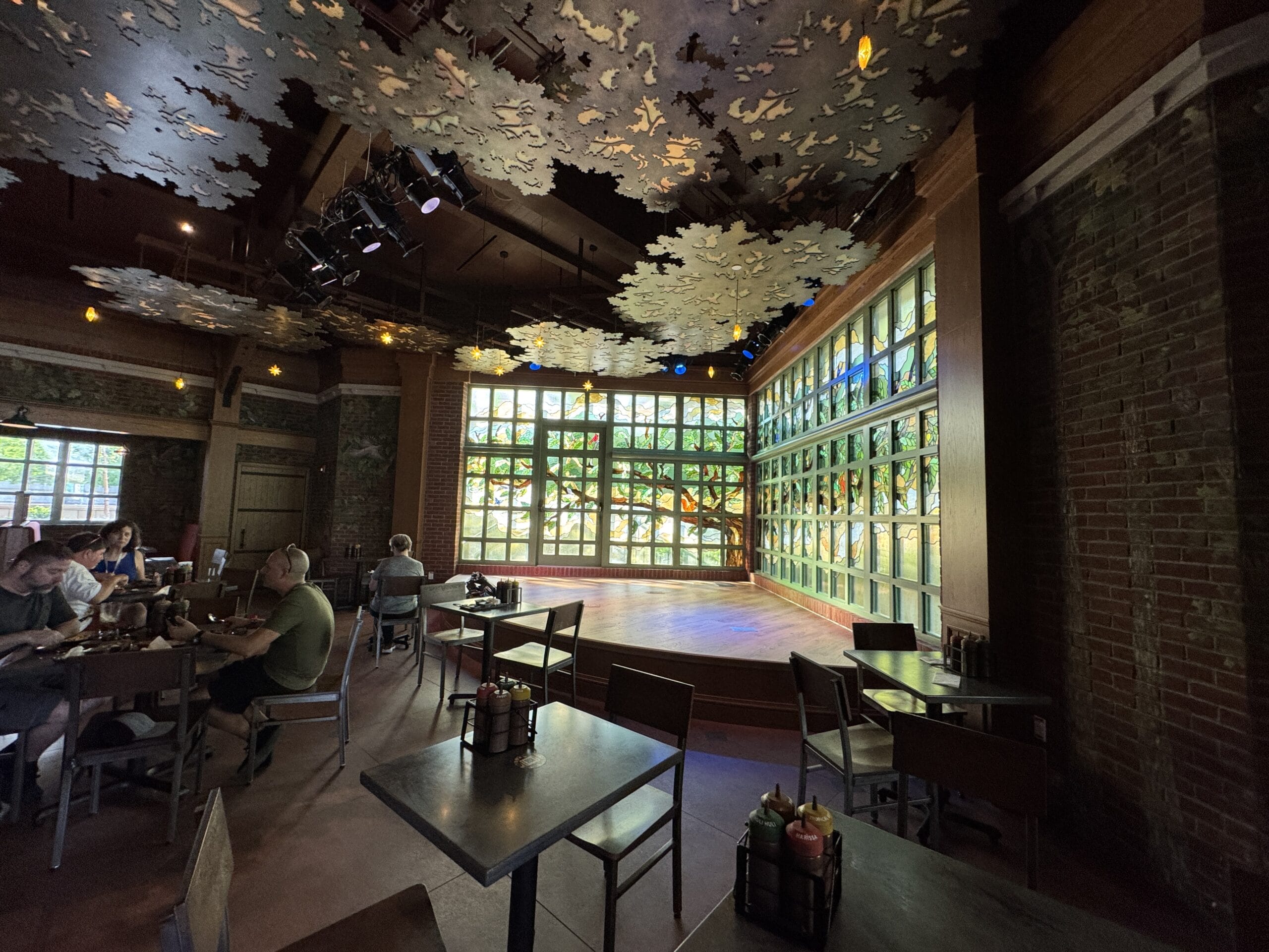 Spacious Disney park restaurant, The Oak & Star Tavern, large windows, empty stage, diners beneath whimsical leafy ceiling decor.