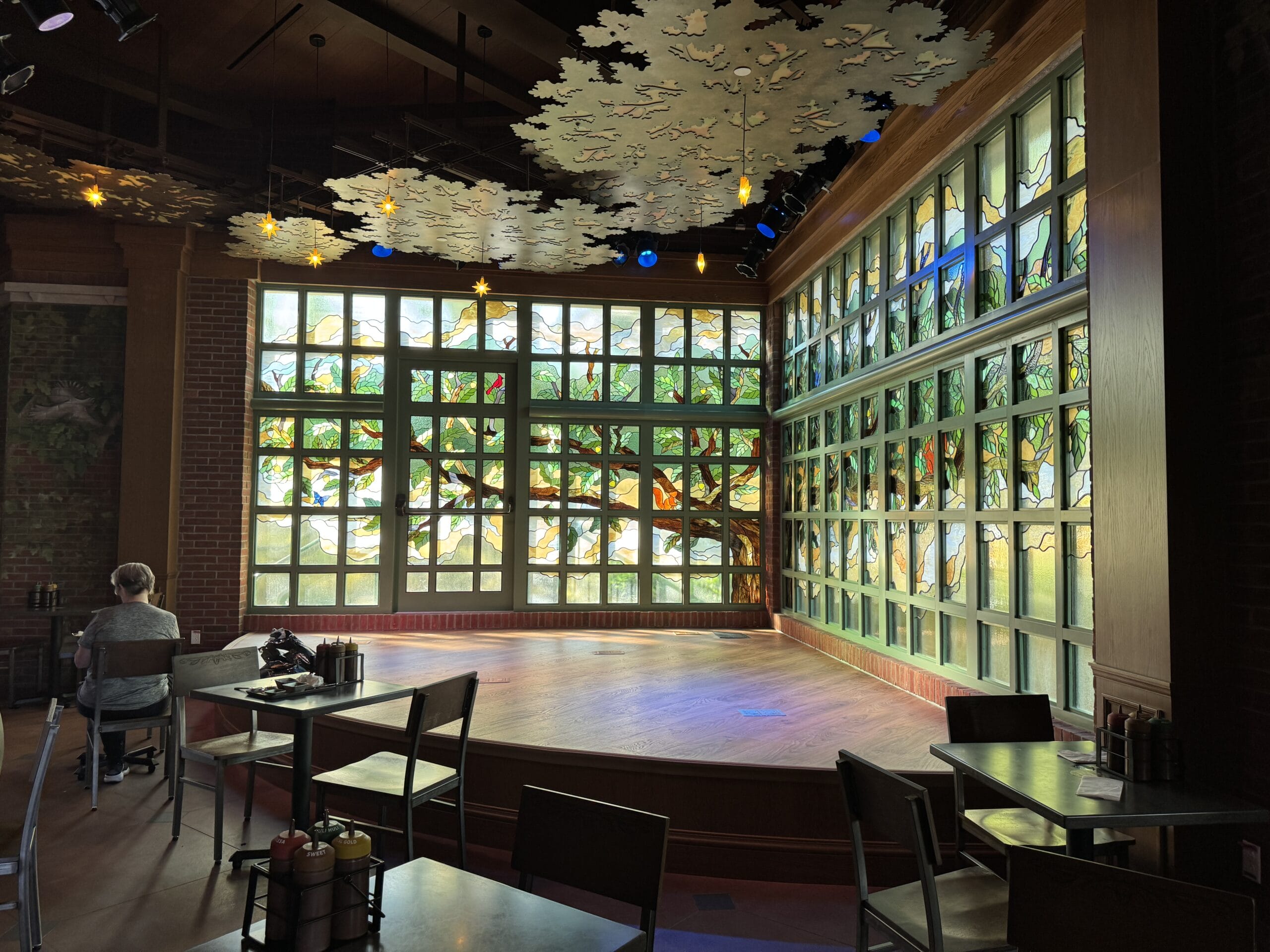 A cozy stage at The Oak & Star Tavern, with stained glass windows recalling Fantasyland vibes, tables, and one guest seated alone.