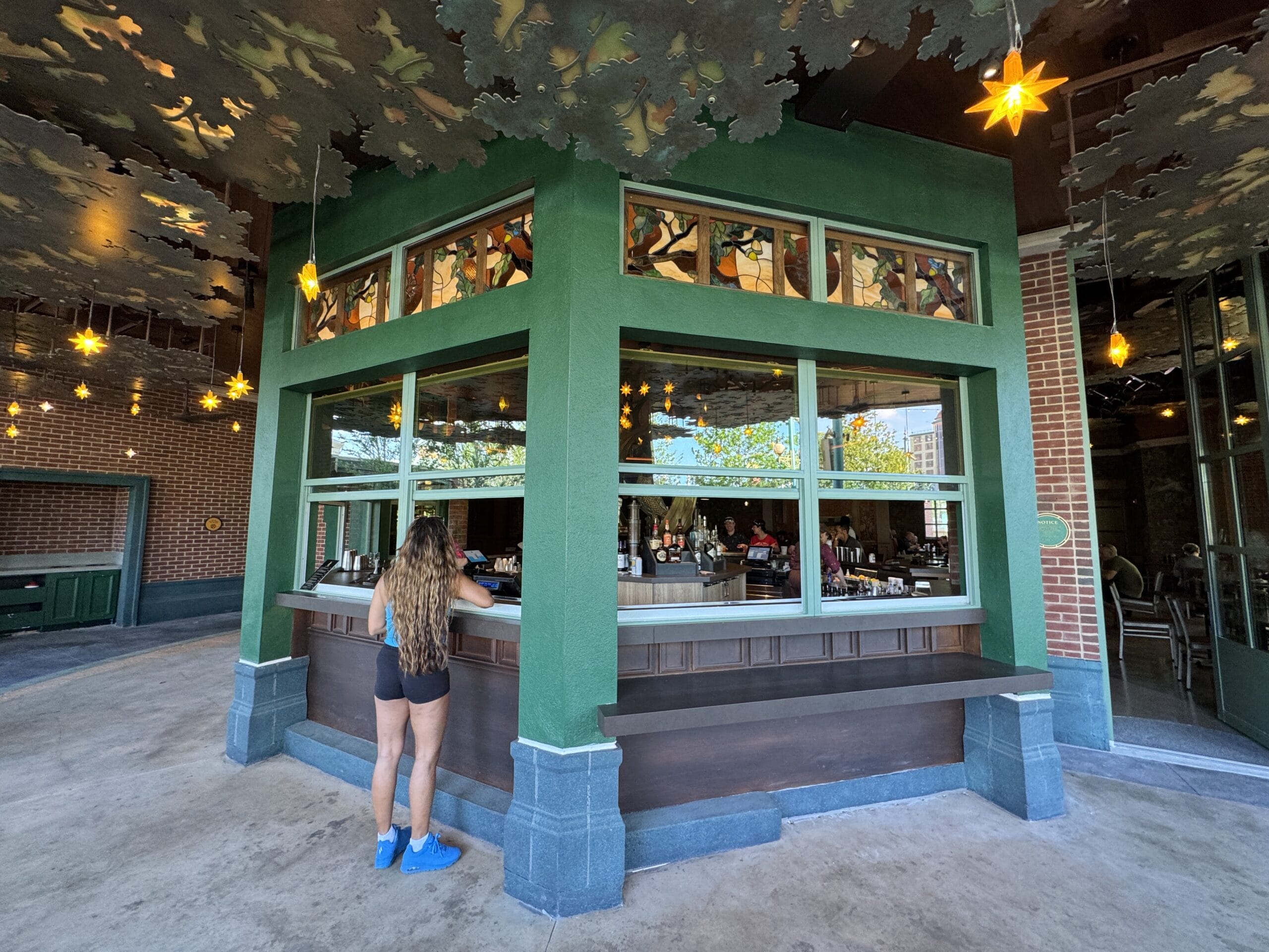 A woman stands at The Oak & Star Tavern’s outdoor bar, lit by hanging lights and ceiling panels, a setting unlike Disney castles.