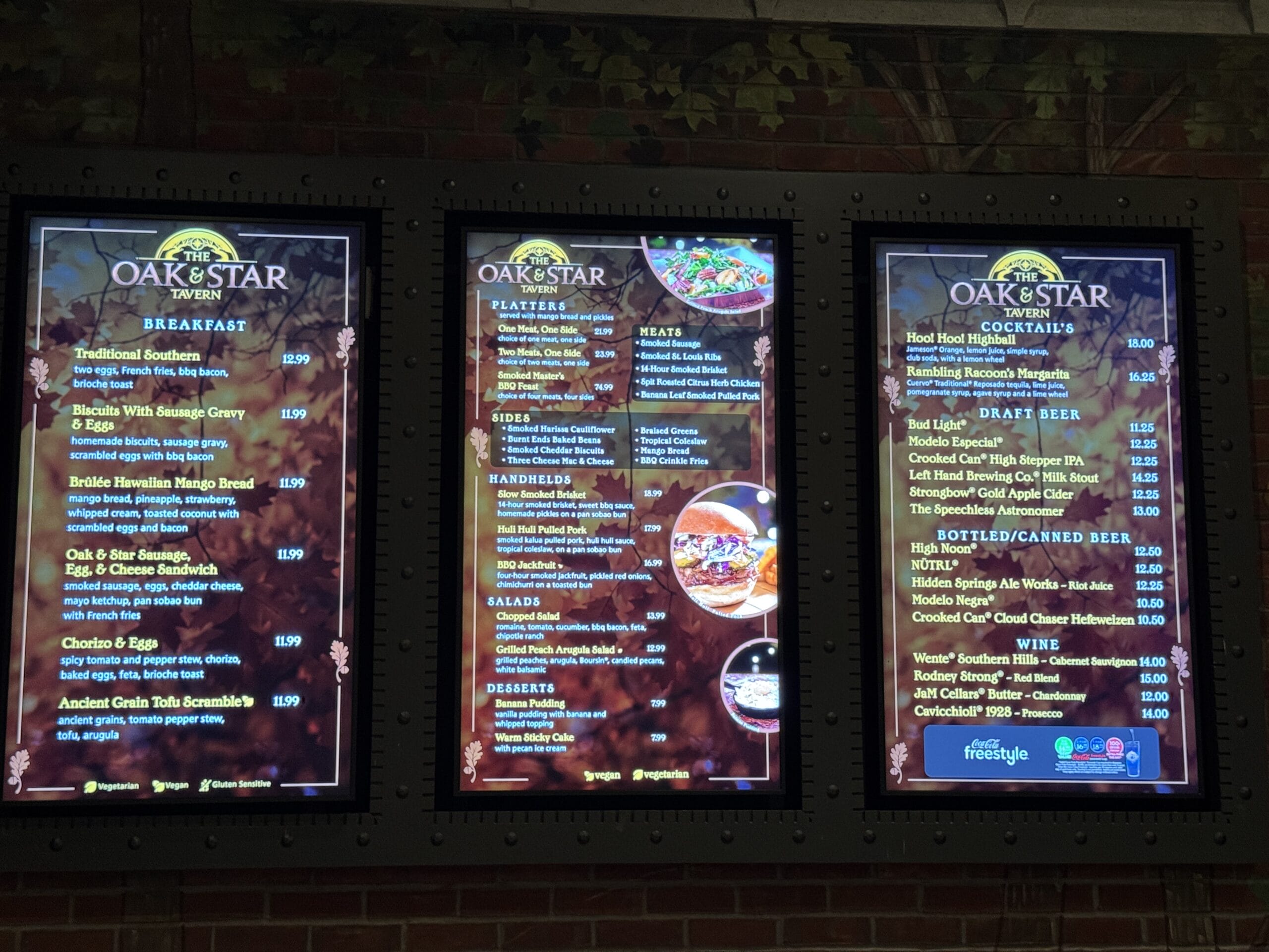 Three digital menu boards at The Oak & Star Tavern in Disney park show breakfast, meats, sides, salads, drinks, and prices.