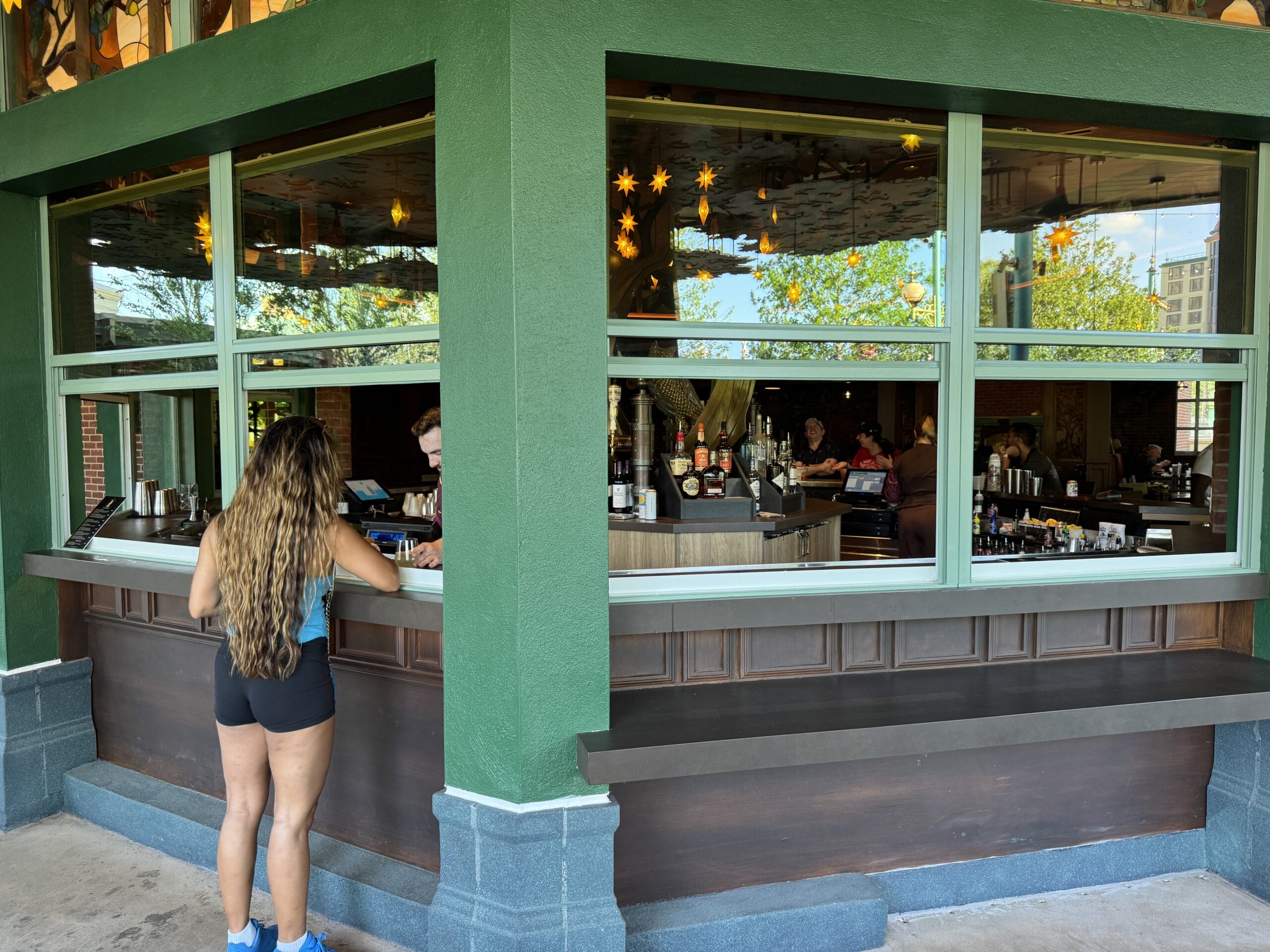 Woman with long hair orders at The Oak & Star Tavern café window; inside, guests mingle under star-shaped lights like Fantasyland décor.