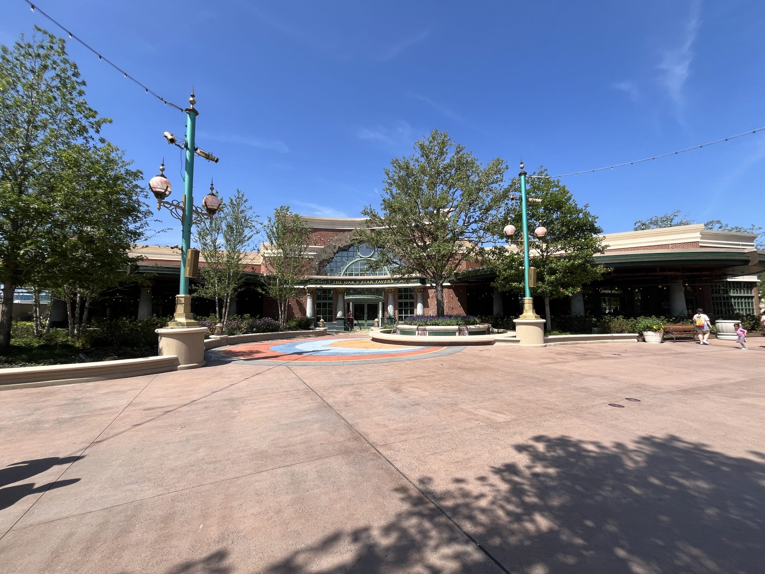The Oak & Star Tavern, a large building with green trim, stands by trees and a vibrant plaza, evoking Disney park theming.