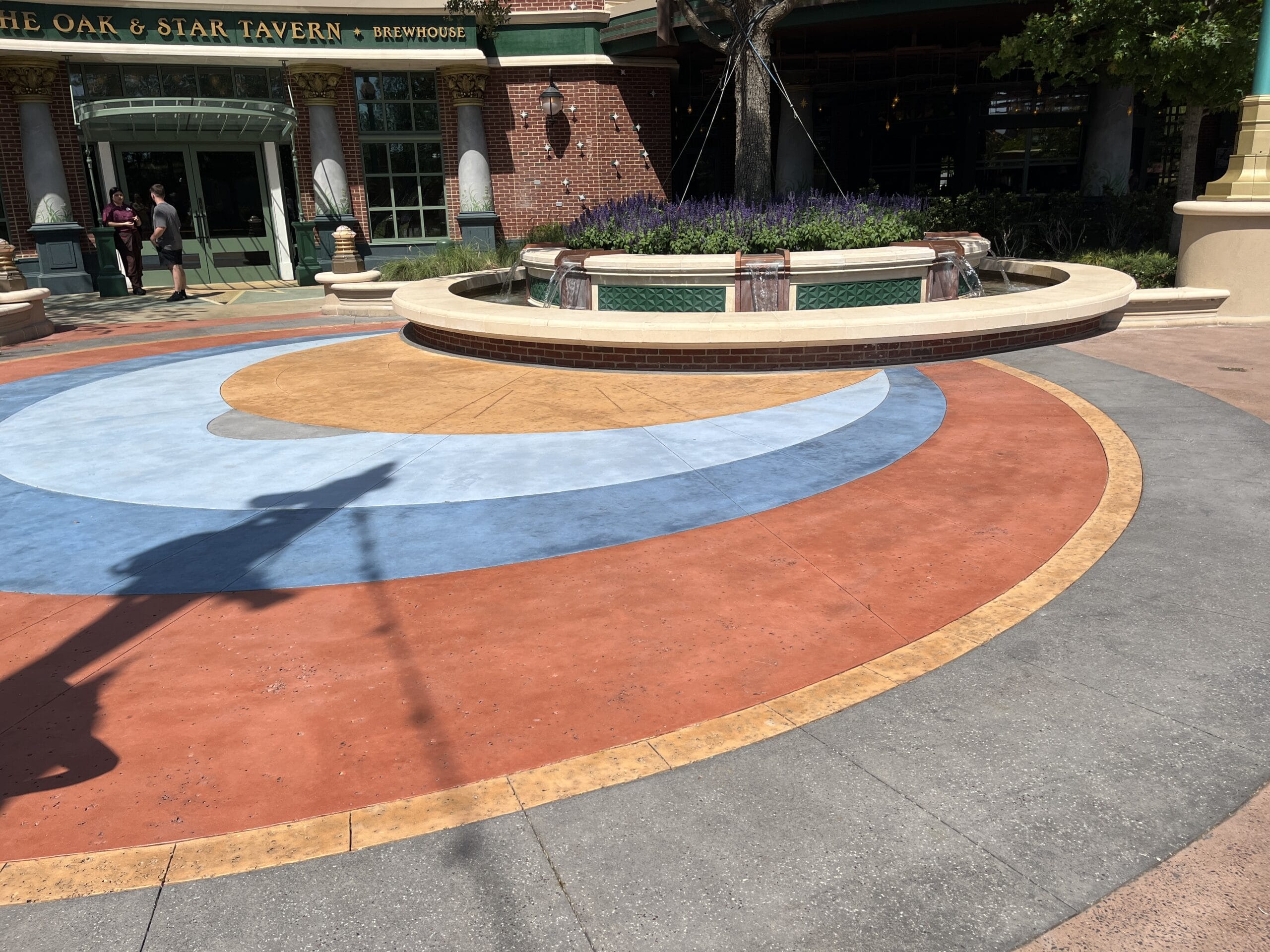 A circular fountain with patterned pavement sparkles outside The Oak & Star Tavern, reminiscent of Disney park entry plazas on a sunny day.