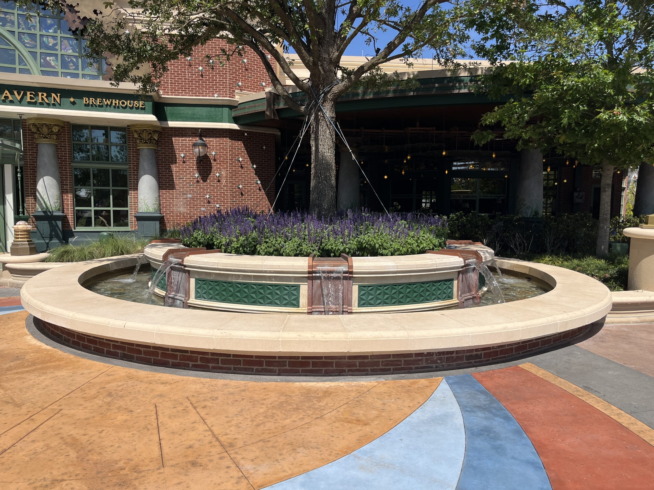 Round stone fountain with water, lavender, and a tree at The Oak & Star Tavern’s entrance; no Disney castle in view.