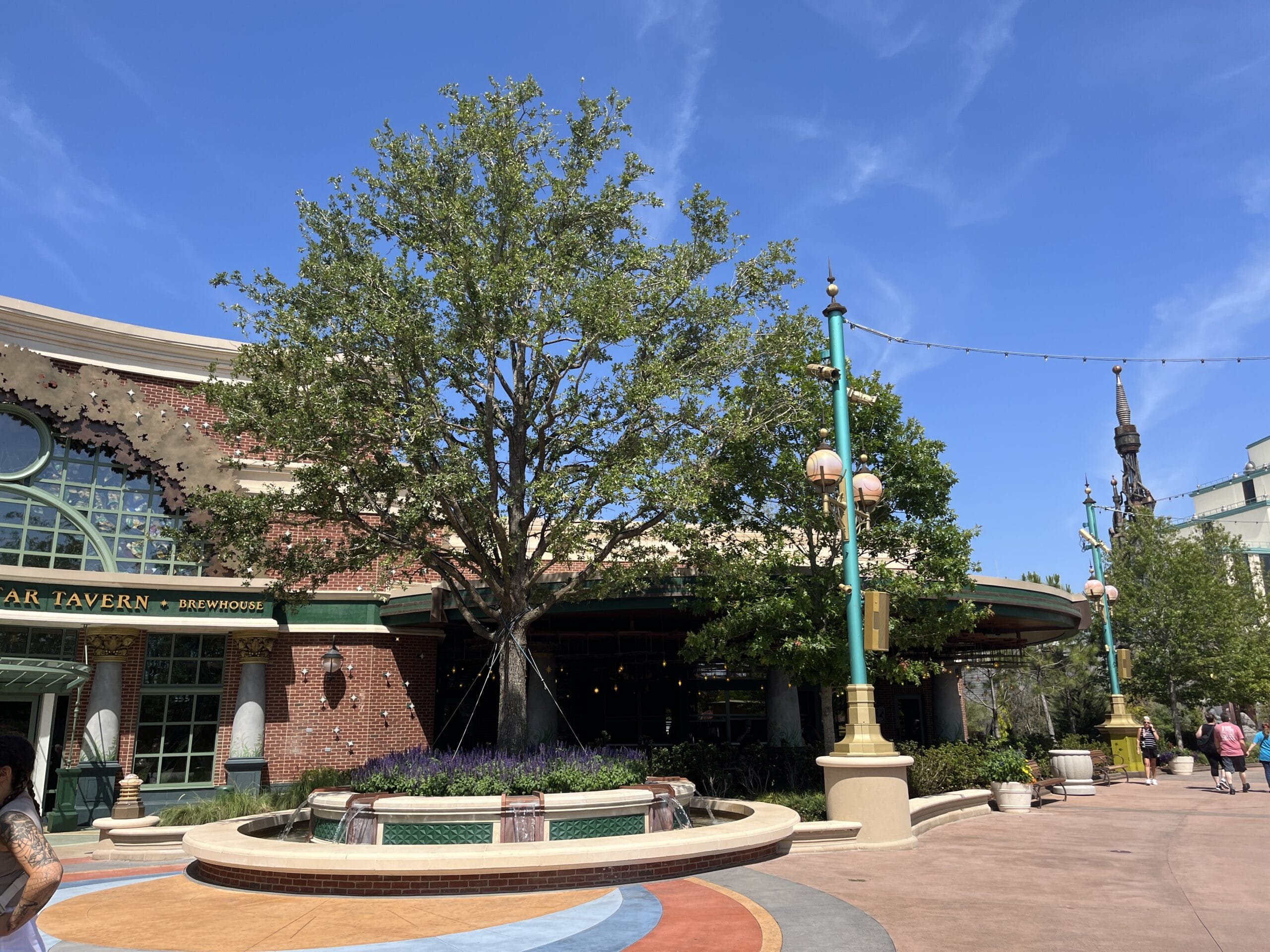 A tree stands before The Oak & Star Tavern, its green-trimmed brick facade beneath a blue sky, echoing Main Street’s charm at Disney parks.