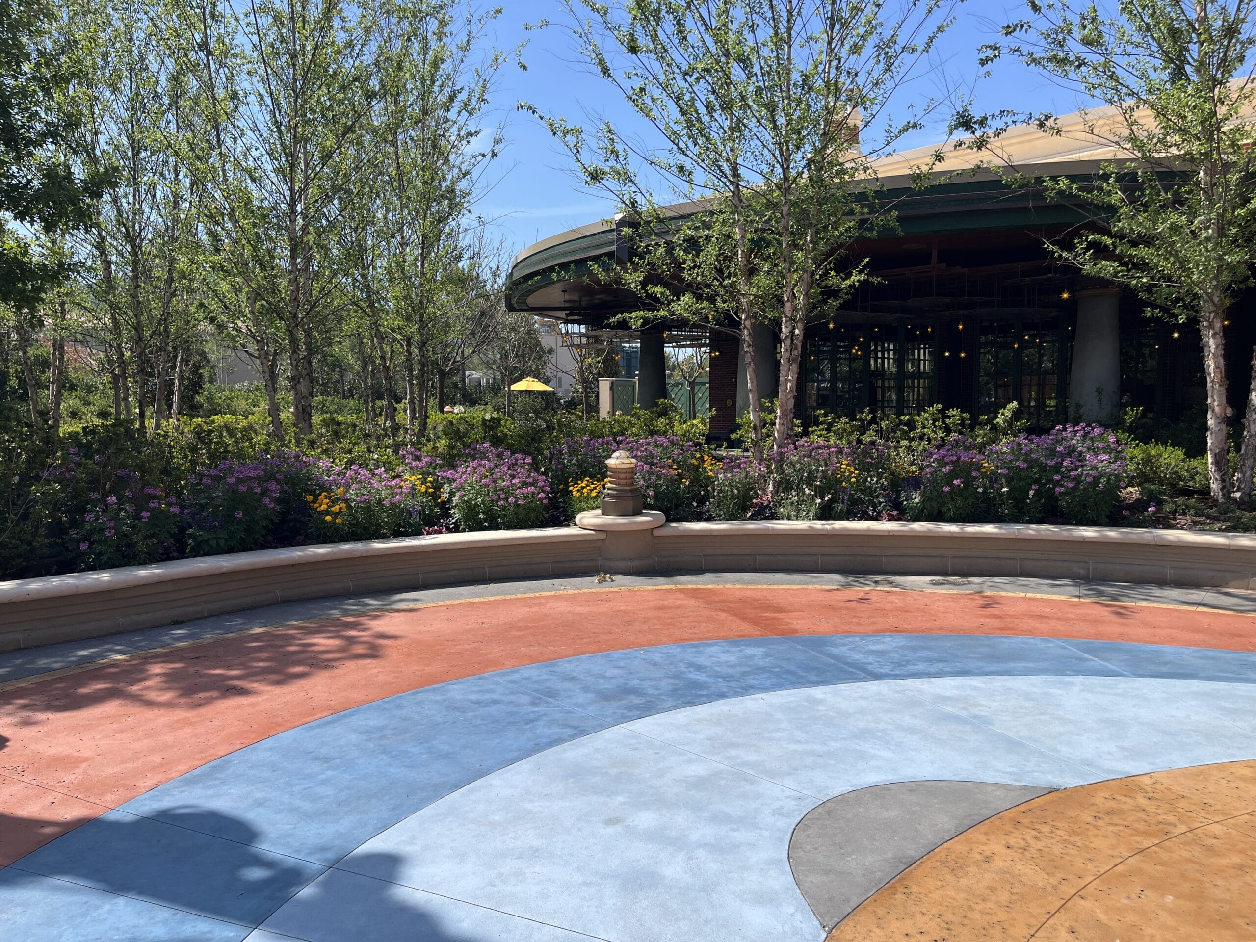 Curved benches and a small fountain by The Oak & Star Tavern, amid vibrant paving, flowers, and trees in a Disney park setting.