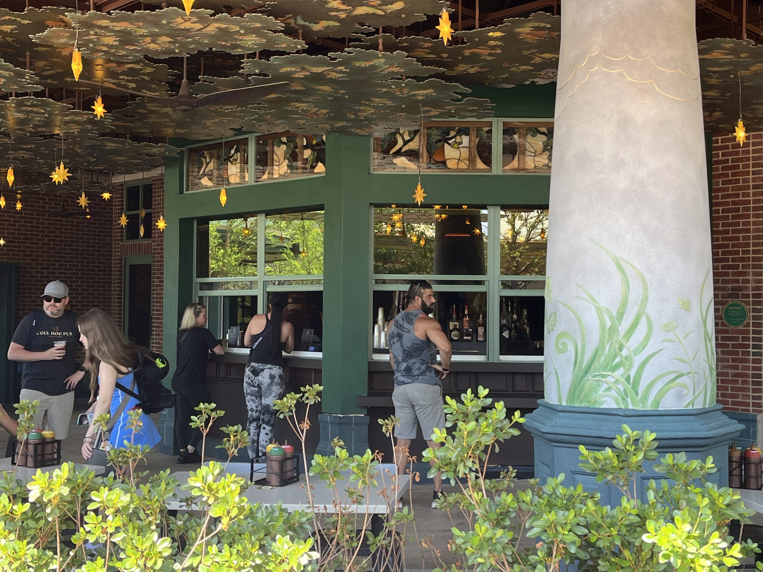 Guests line up at The Oak & Star Tavern’s outdoor counter, framed by green trim, glowing lights, and foliage—no castle in sight.