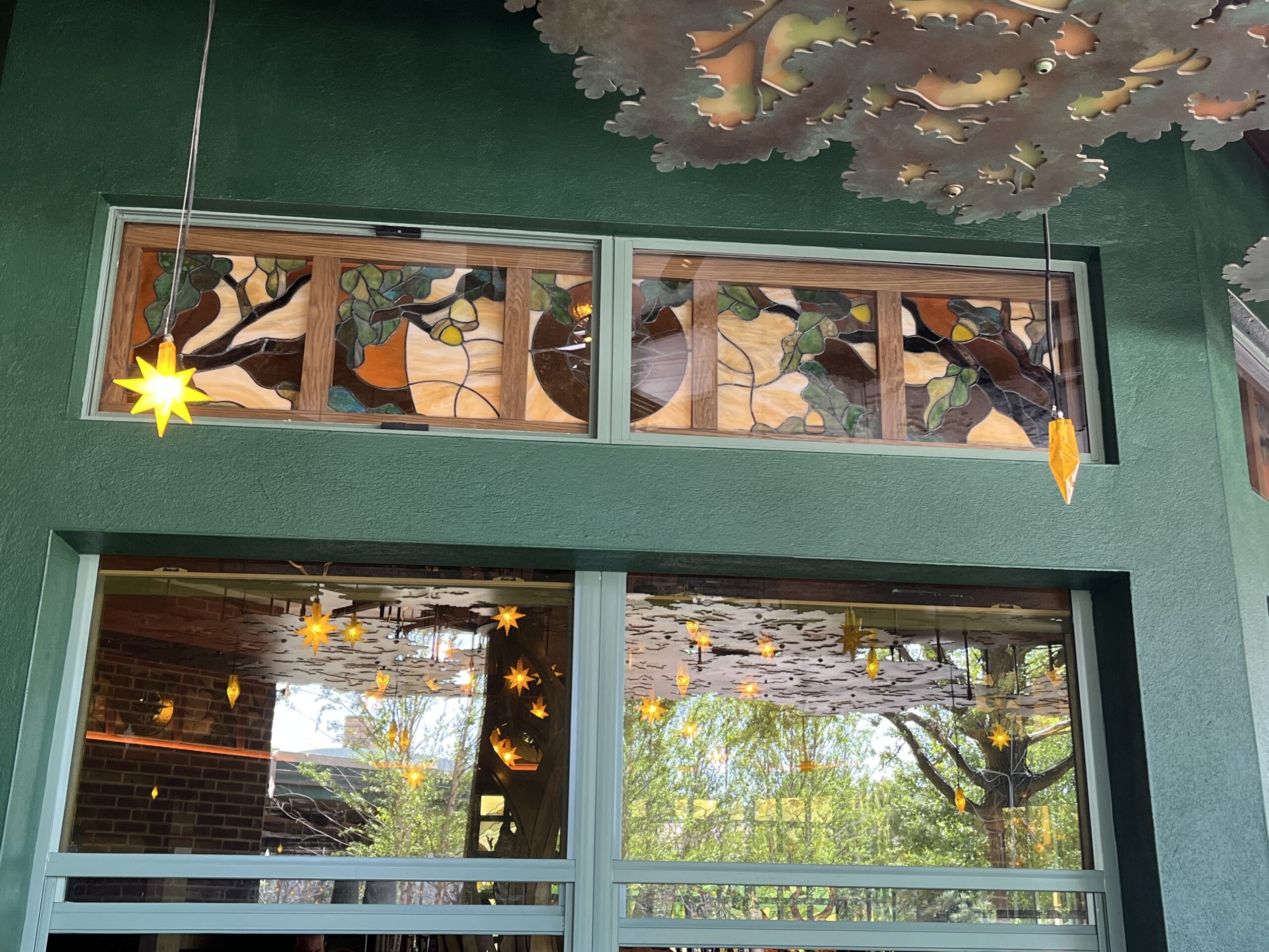 Stained glass window with birds and leaves above The Oak & Star Tavern’s doors, star-shaped lights hang below—evokes Fantasyland charm.