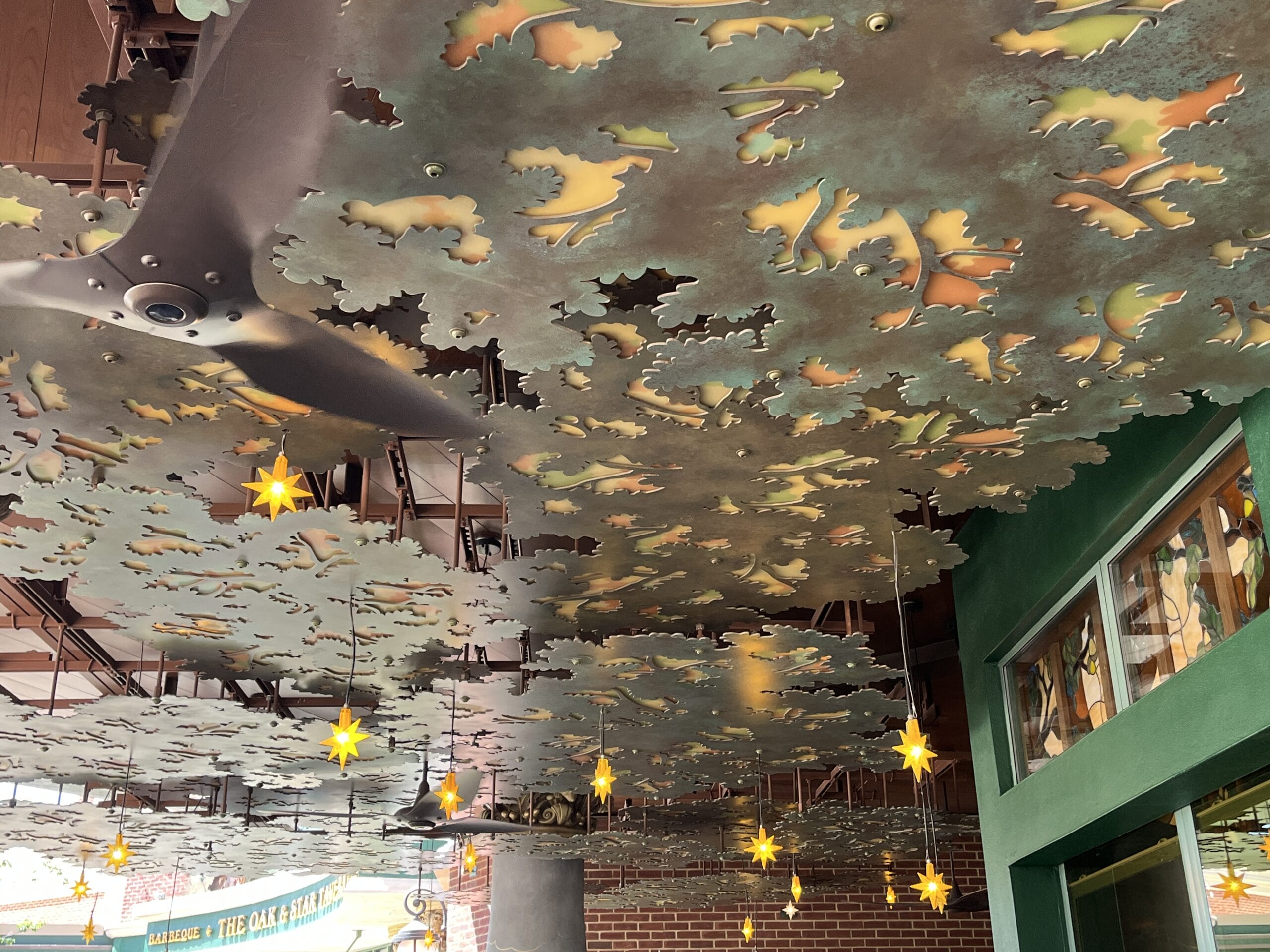 Ornate metal ceiling panels and star lights glow above The Oak & Star Tavern’s leafy wall, evoking a Disney park’s enchanting decor.
