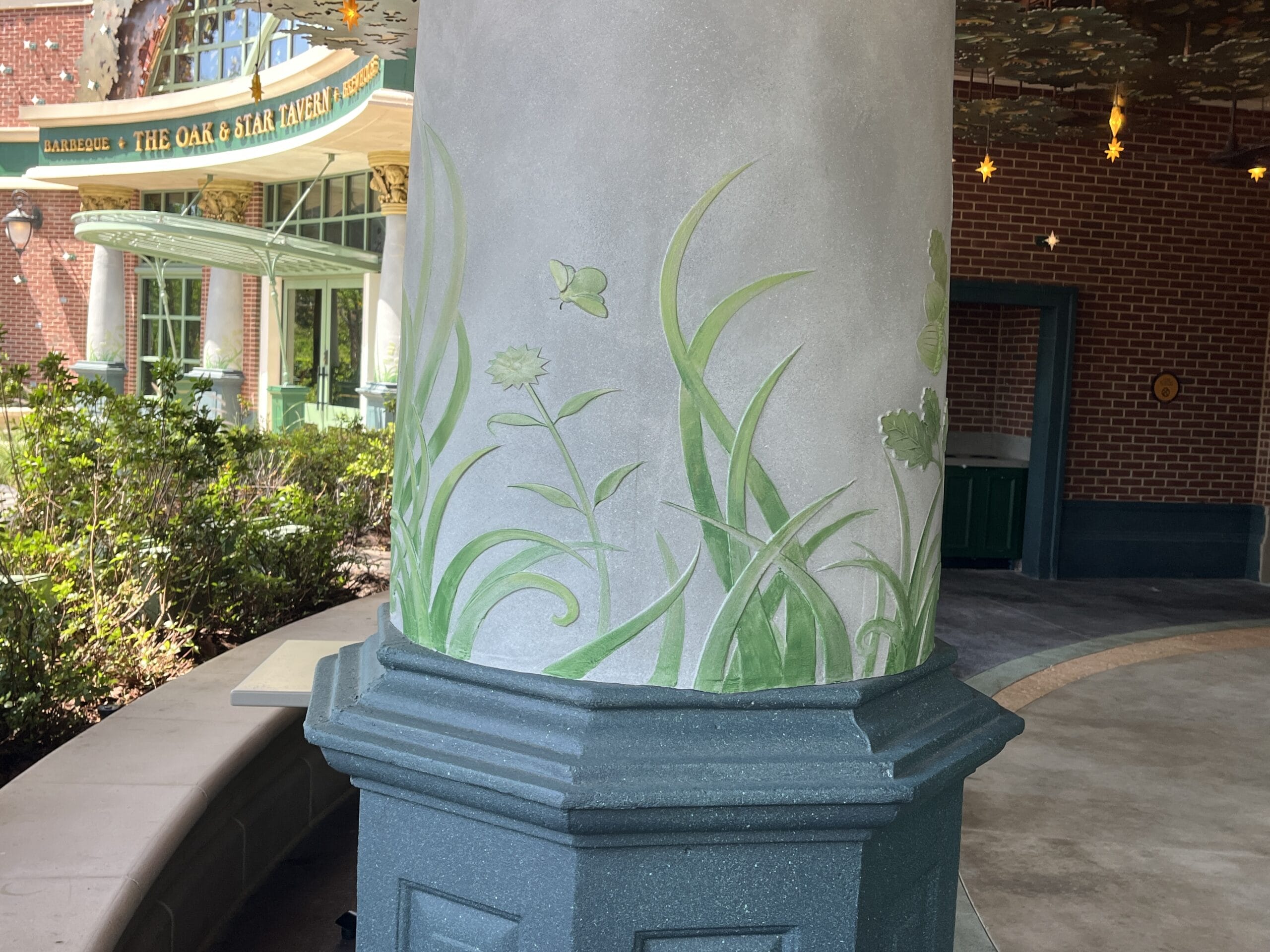 A painted pillar with green grass, plants, and insects at The Oak & Star Tavern entrance, sign above—no Disney castles here.