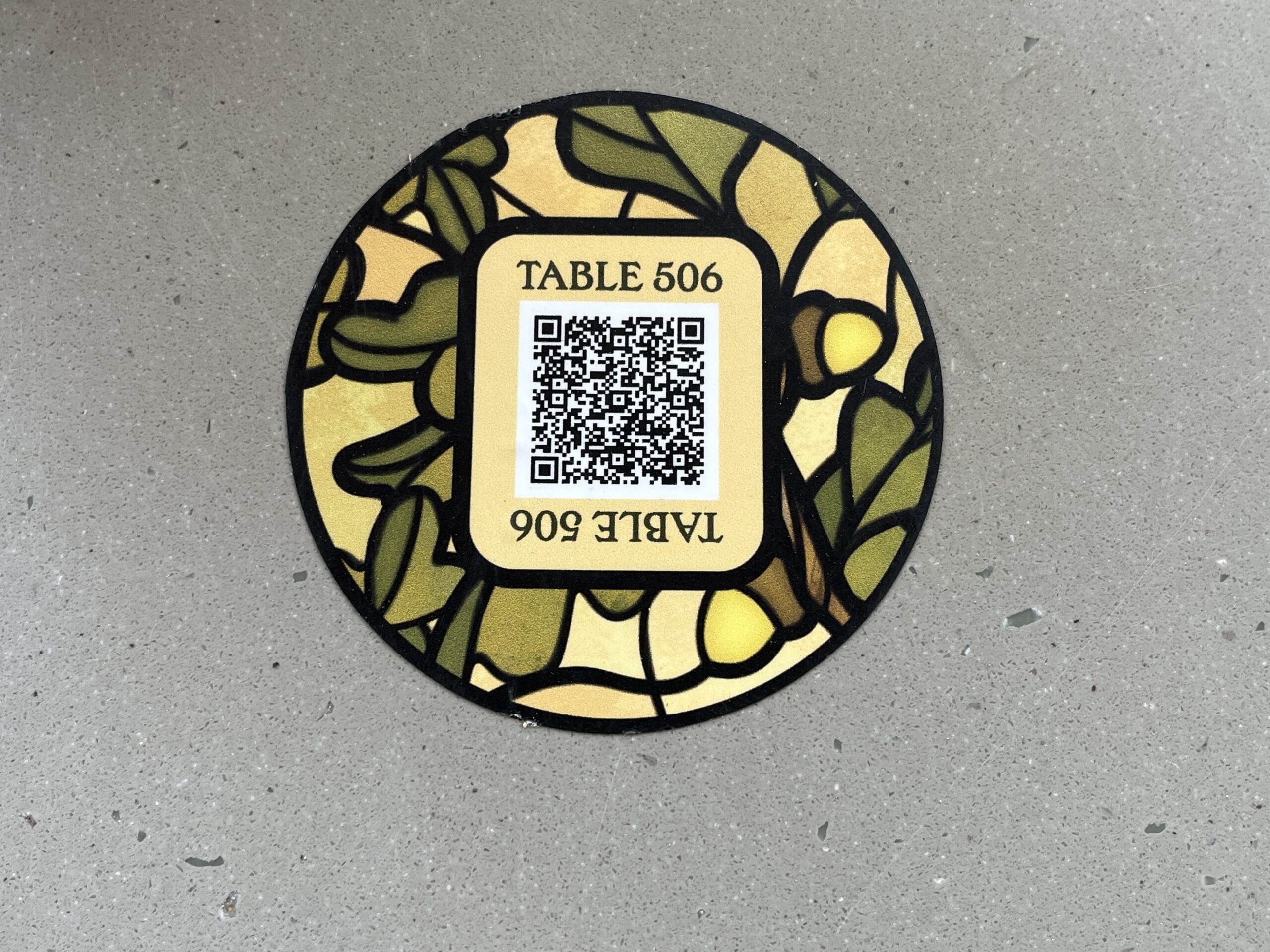A stained-glass style coaster from The Oak & Star Tavern with a QR code and “TABLE 506,” shown on a gray speckled tabletop; not a castle like any at Disney Parks.