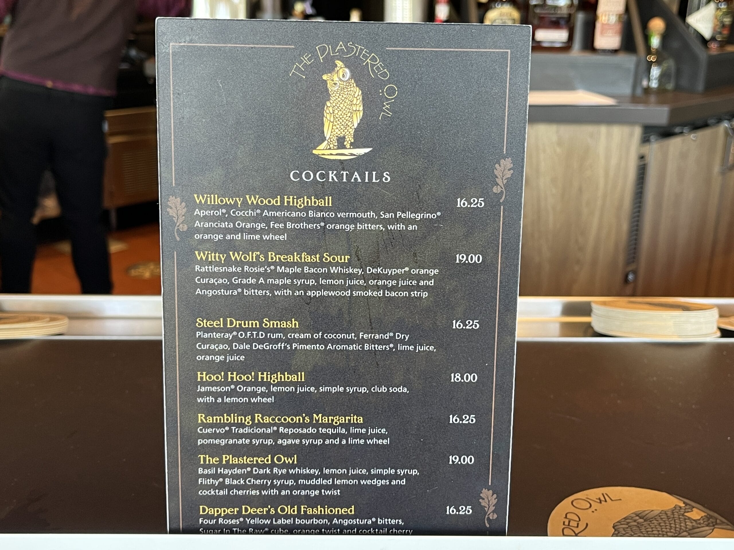A themed cocktail menu from The Plastered Owl and Oak & Star Tavern sits on a bar, listing drinks and prices like in Disney parks.