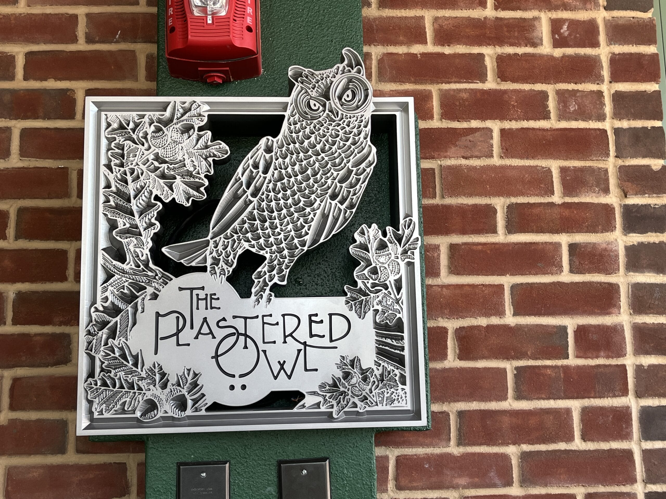 A themed metal sign featuring an owl and "The Plastered Owl" hangs on a brick wall just outside The Oak & Star Tavern, evoking Disney park detail.