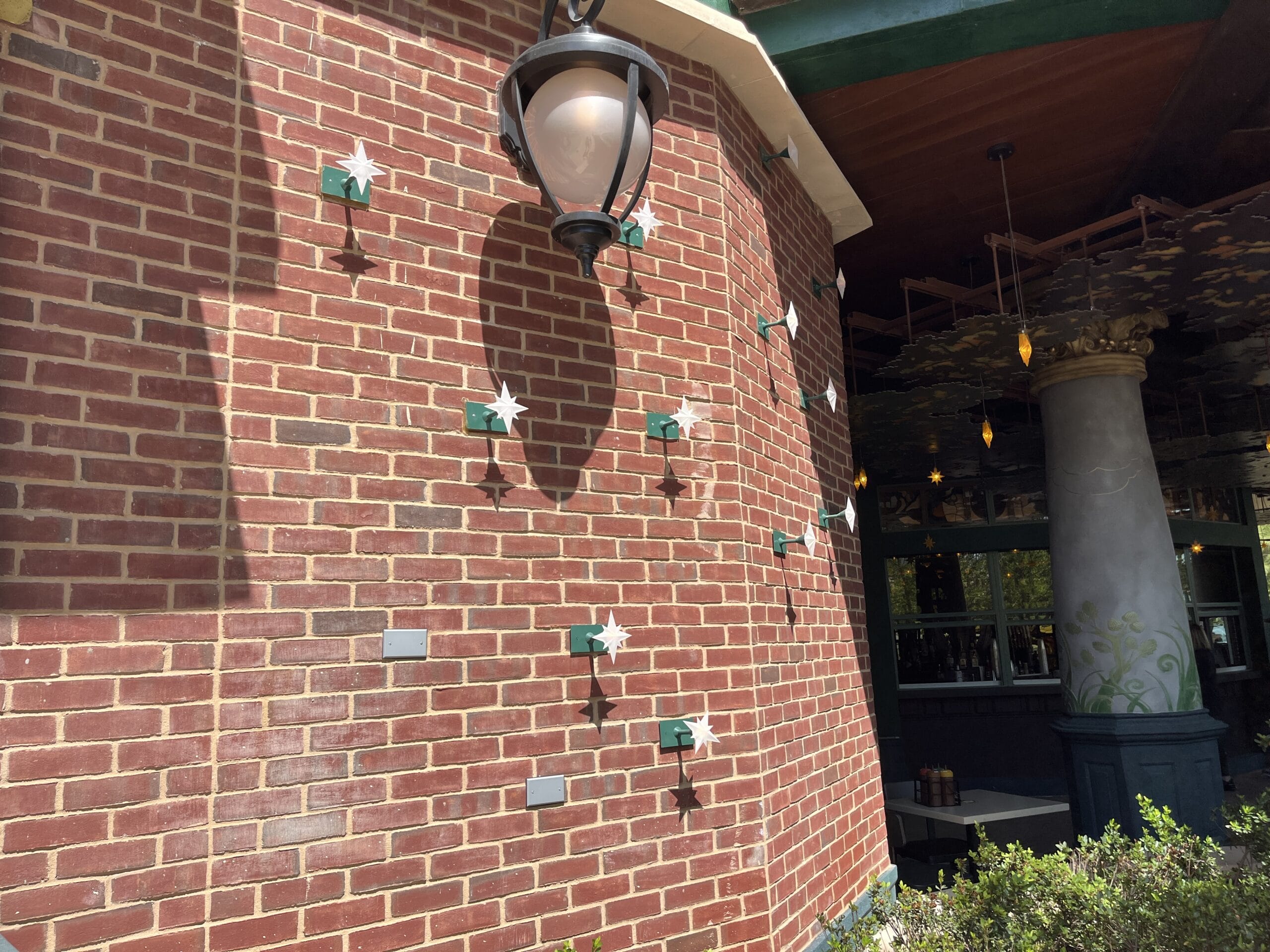Brick wall at The Oak & Star Tavern with green and white flower sculptures, plaques, and patio lantern, no Disney castle present.