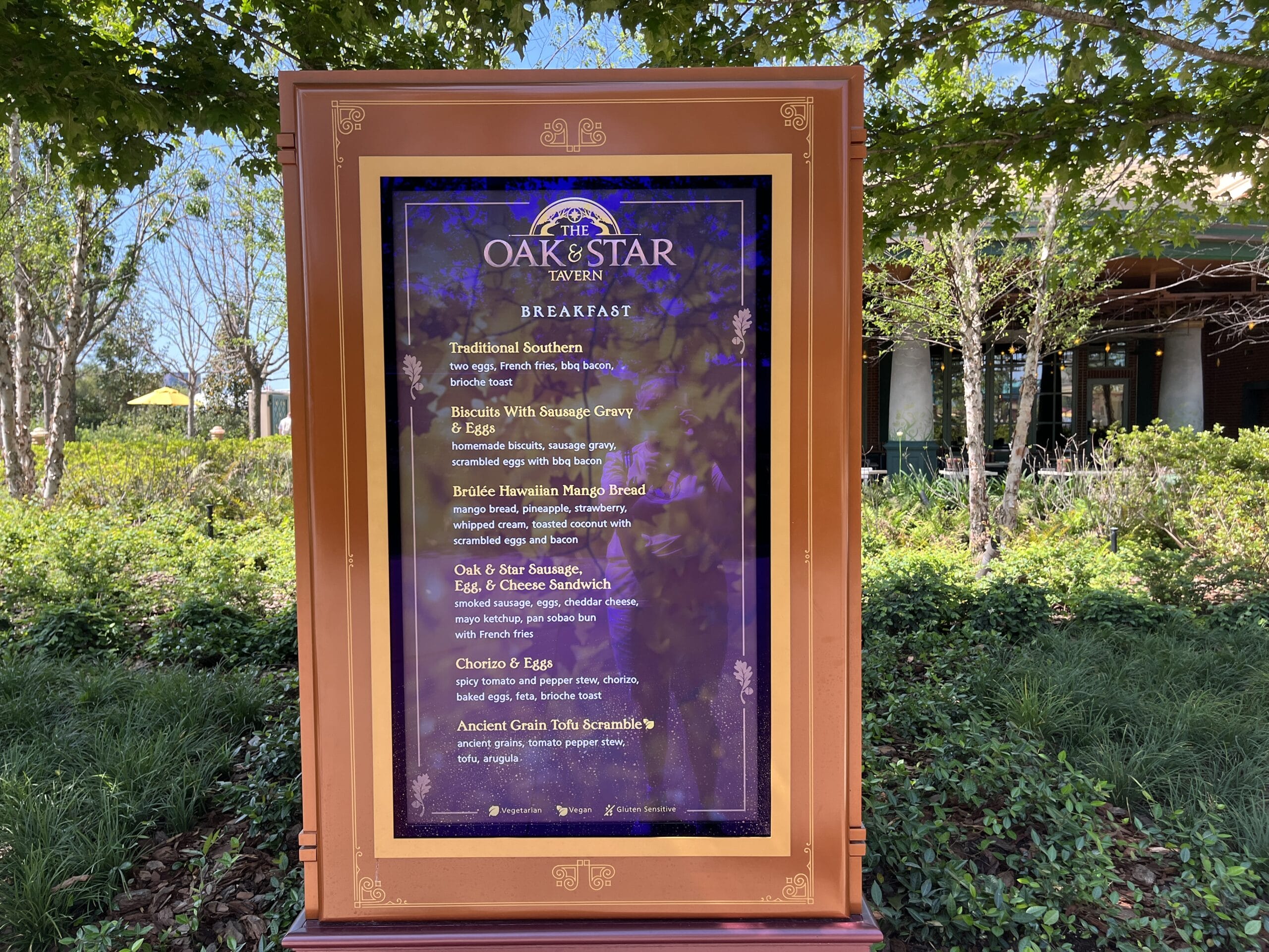 The Oak & Star Tavern breakfast menu stands outside among trees in a Disney park, listing morning meals near classic Disney theming.