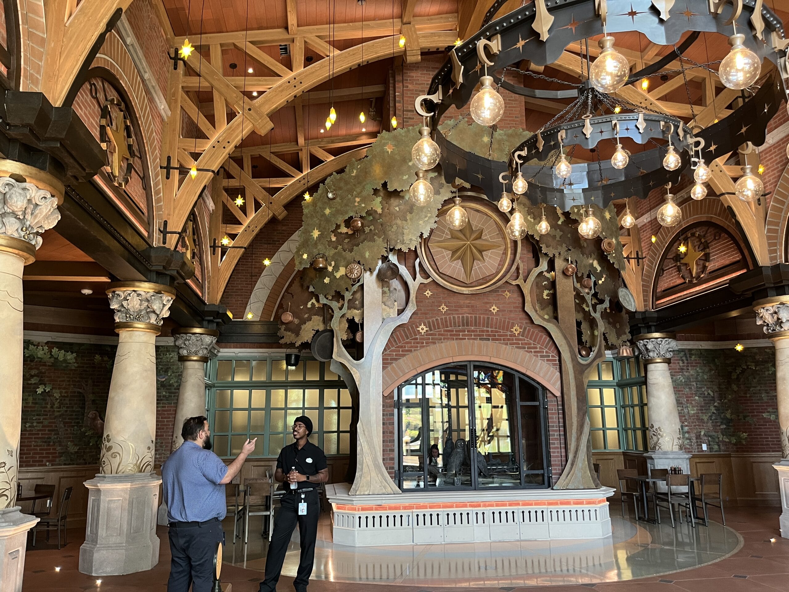 Two people chat inside The Oak & Star Tavern, a lavish hall with columns, chandeliers, and arches—evoking Disney park elegance.