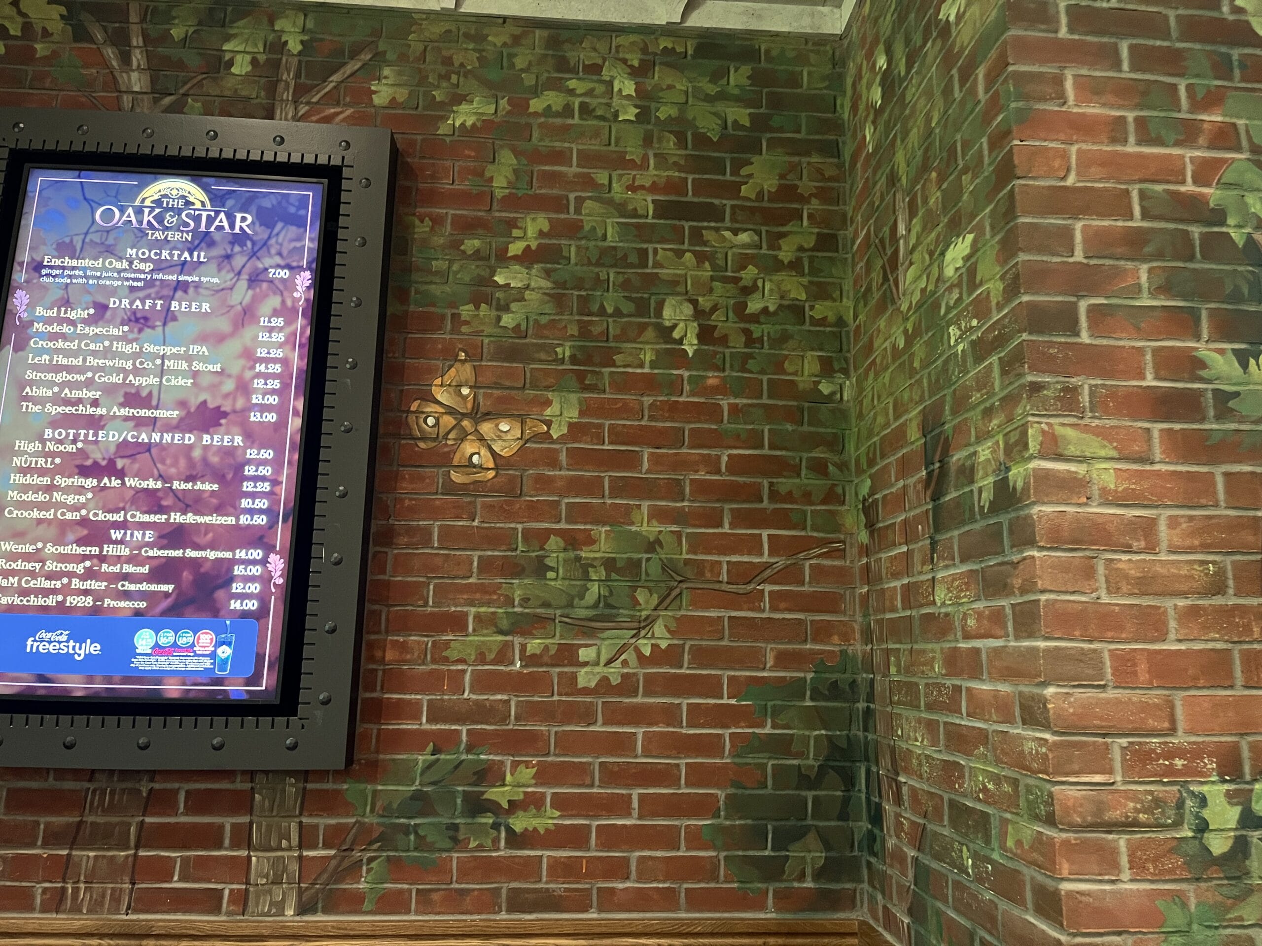 A digital menu board at The Oak & Star Tavern on a brick wall, with green vine decor and a butterfly—reminiscent of Disney parks' themed touches.