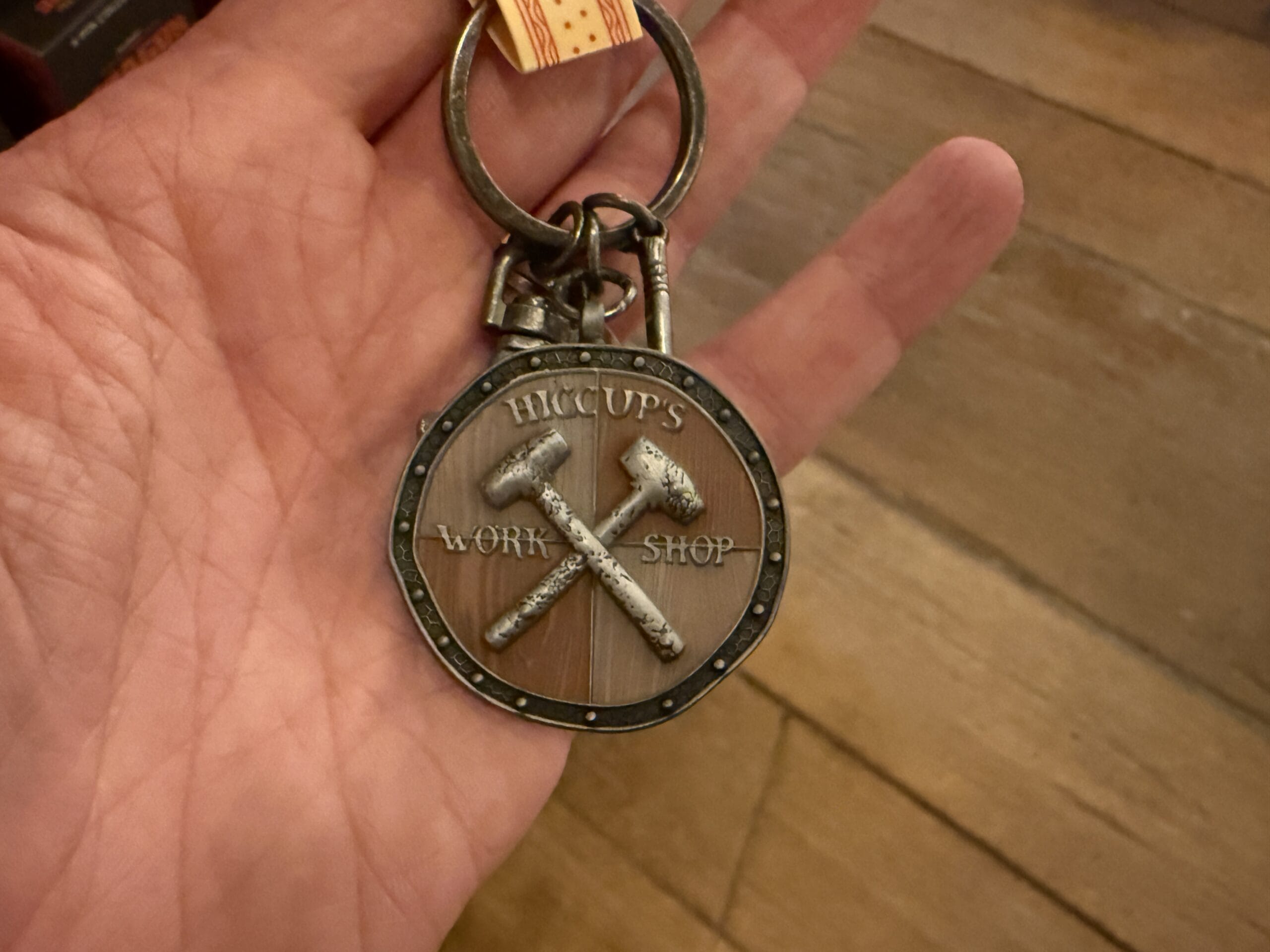 A hand holds a Viking Traders keychain labeled "HICCUP'S WORKSHOP" with crossed hammers on a wooden background.