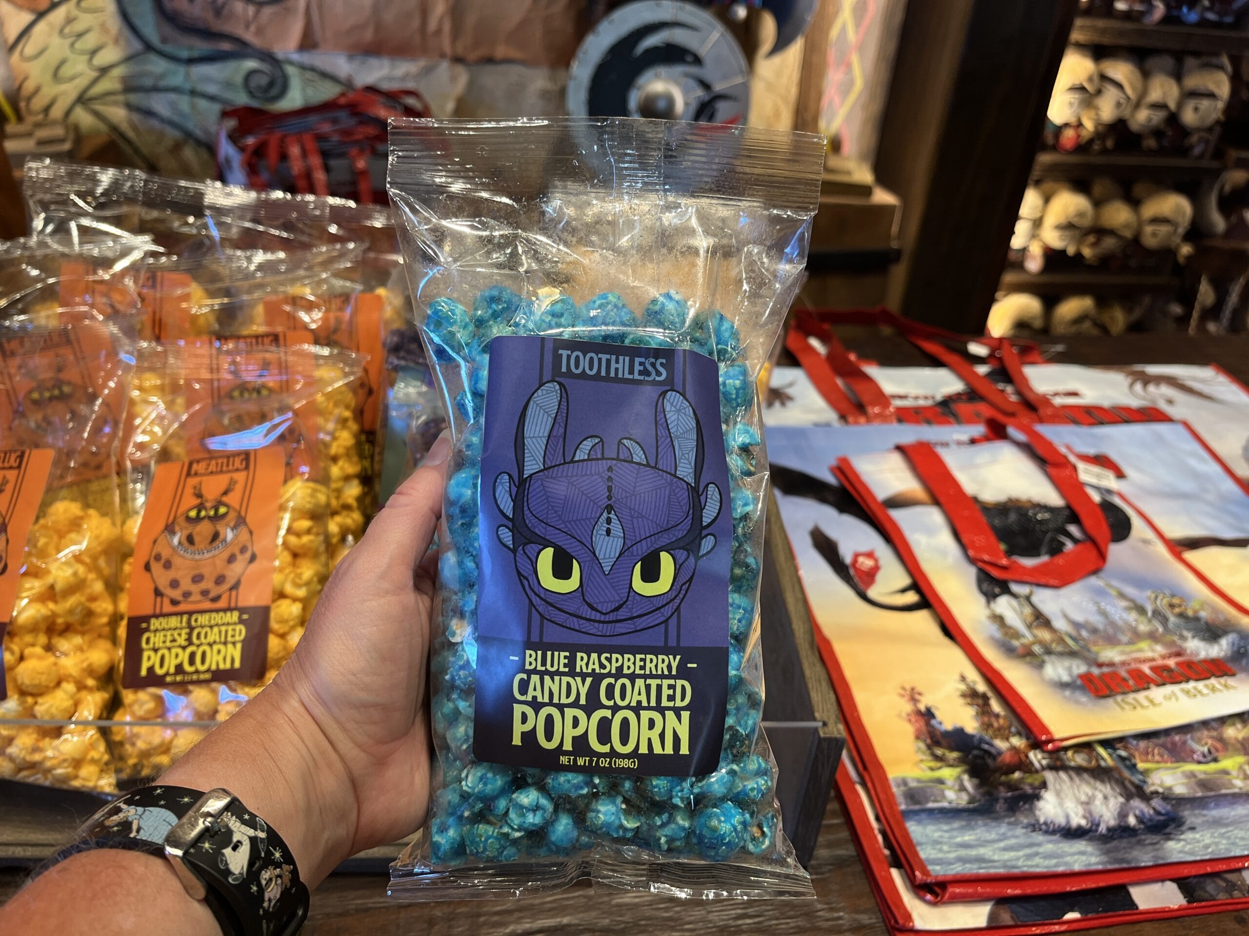 A hand holds a bag of Toothless blue raspberry popcorn from Viking Traders amid other popcorn and red bags in a store.