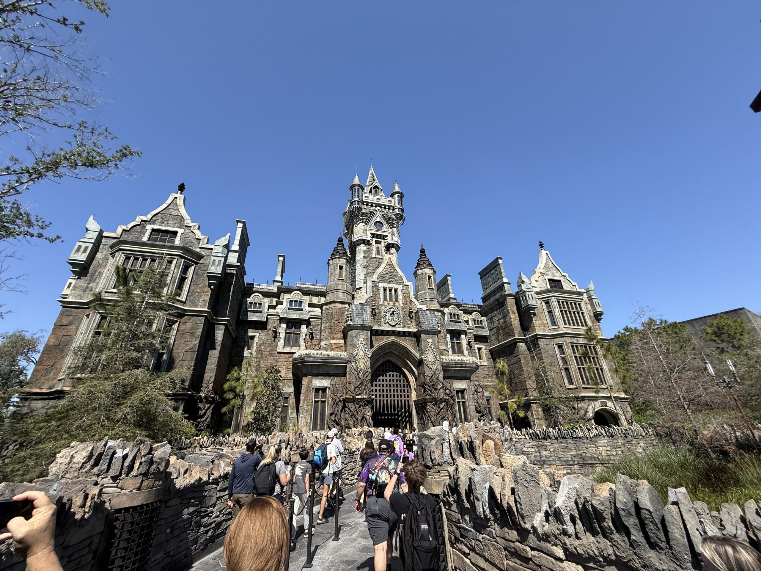 A group of people walk toward a large, gothic-style stone castle with turrets and arched windows under a clear blue sky.